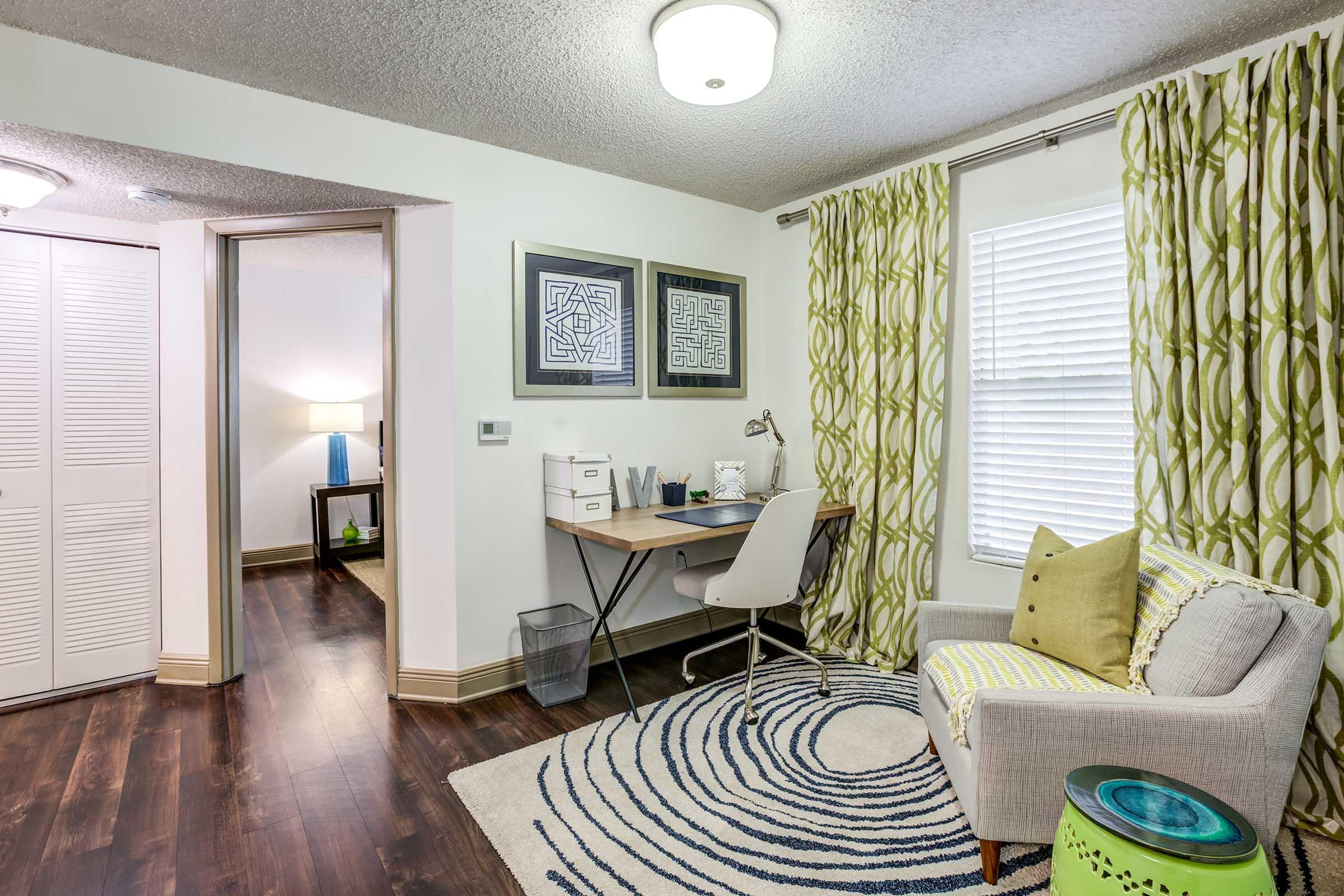A cozy room featuring a desk with a chair, decorative art on the wall, a rug with a spiral pattern, a green accent table, and large windows with patterned curtains. The floors are dark wood, and there's a soft light fixture on the ceiling, creating a welcoming atmosphere.