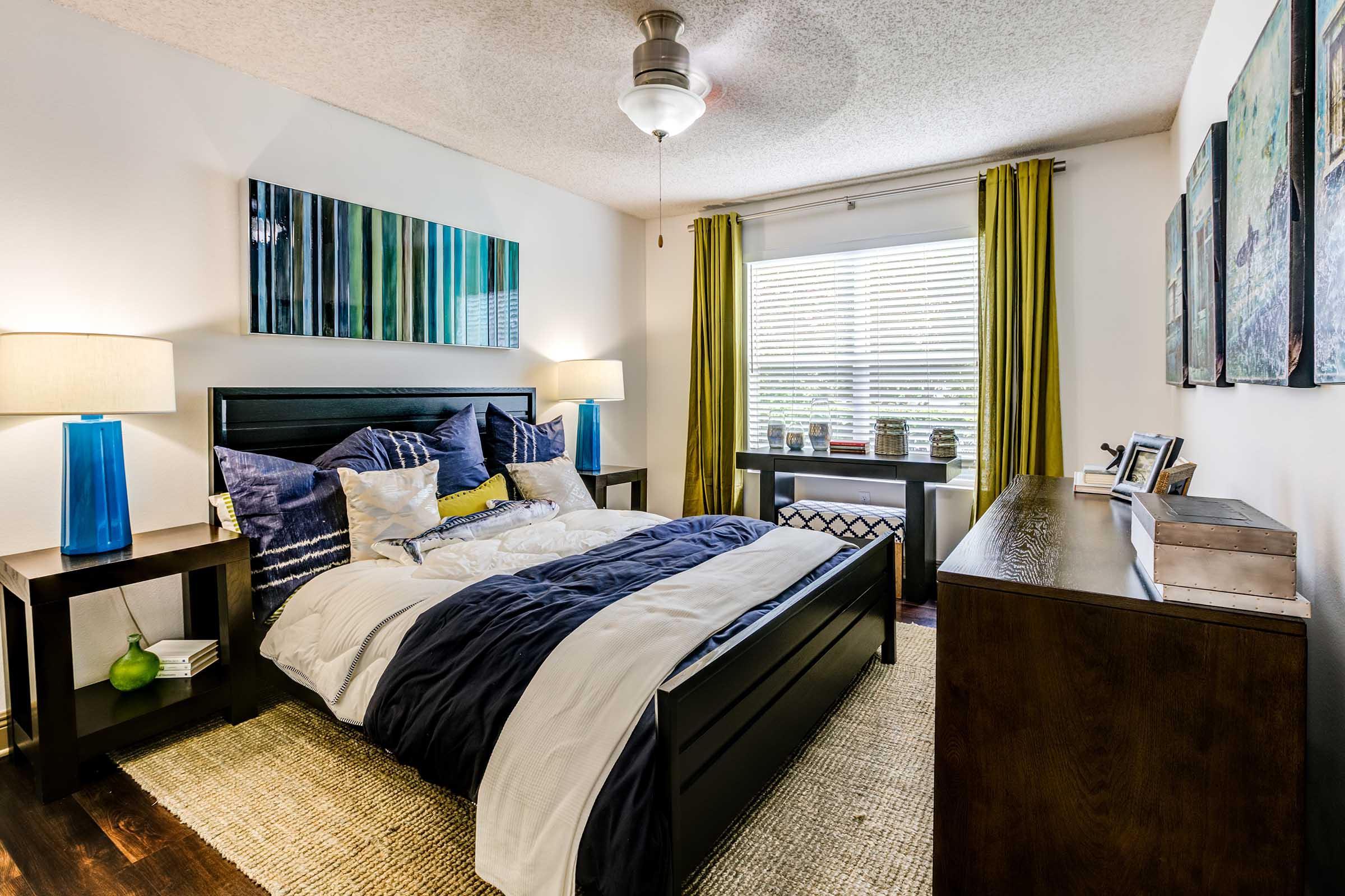 A stylishly decorated bedroom featuring a black bed with blue and white bedding, two bedside lamps with blue bases, and large windows with yellow curtains. A wooden dresser and a window seat adorn the space, complemented by abstract art pieces and decorative items on the window ledge.