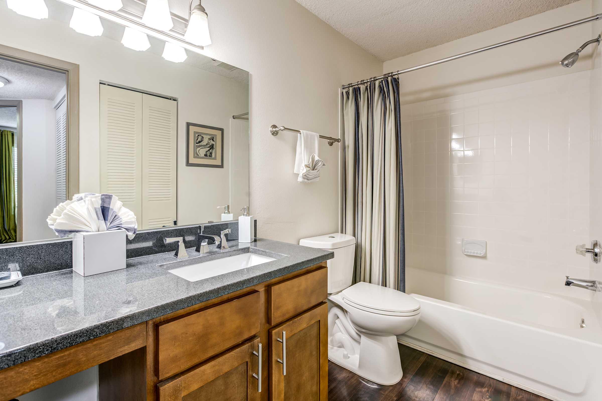 A modern bathroom featuring a double sink vanity with dark wood cabinets, a large mirror, and elegant lighting. There’s a white toilet, a shower/bathtub combo with a curtain, and a decorative towel. The overall color scheme is neutral with touches of dark wood, creating a clean and inviting space.