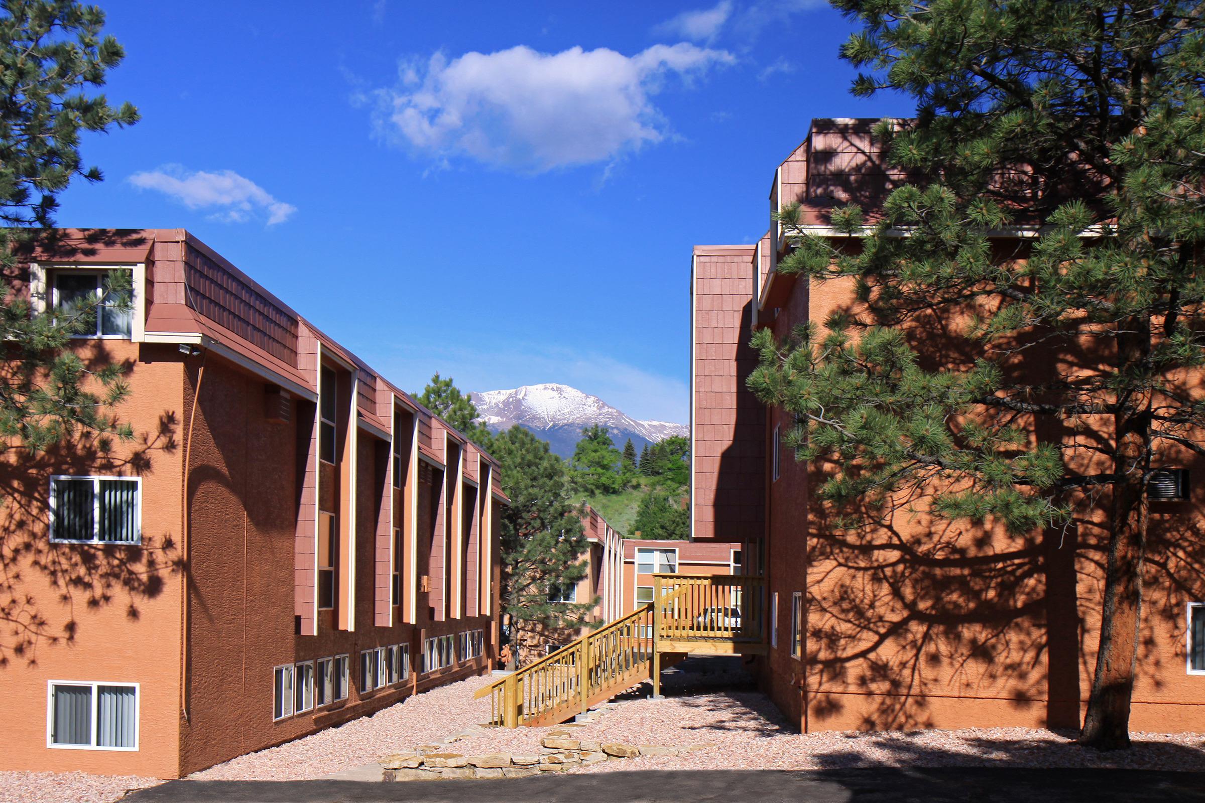 A scenic view of a pathway leading through red-brick buildings, flanked by pine trees. In the background, snow-capped mountains can be seen under a bright blue sky with a few clouds. The image captures a peaceful landscape that combines architecture with natural beauty.