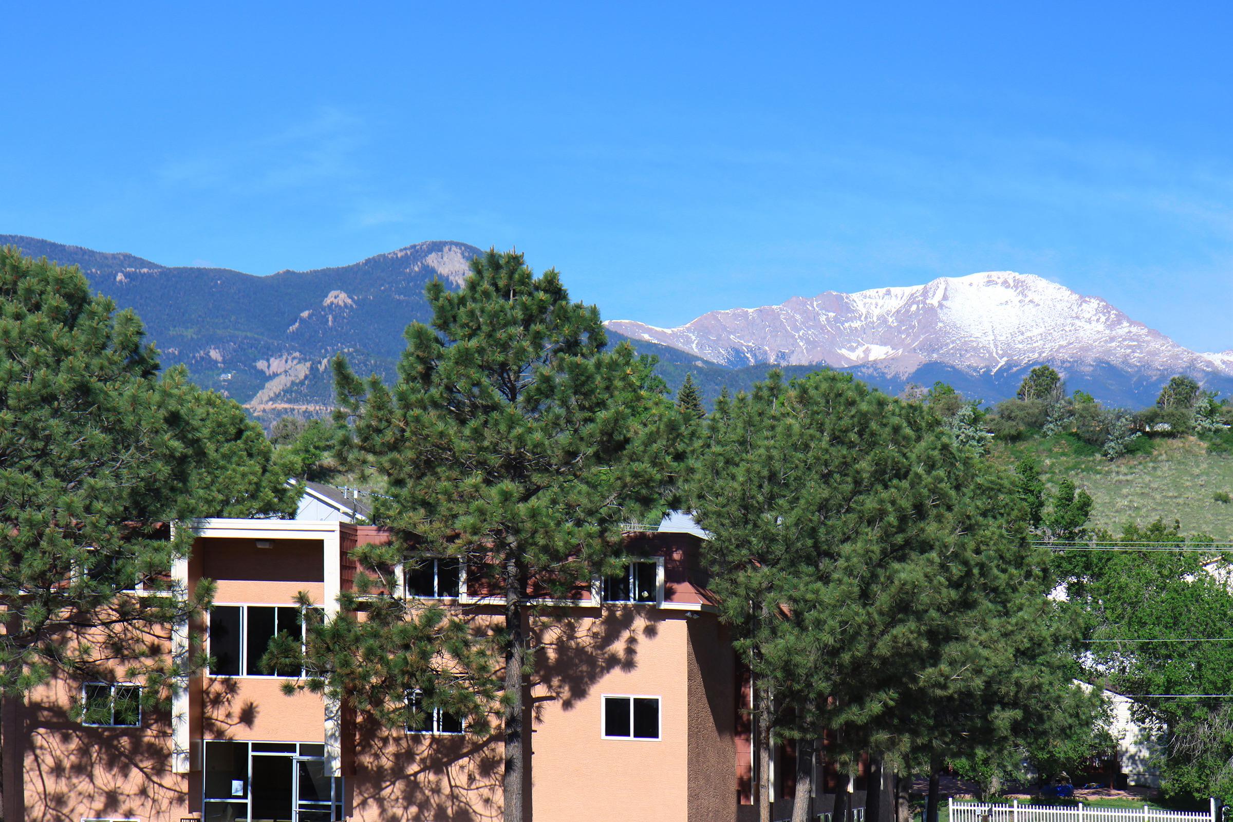 A view of a brown building surrounded by green trees, with snow-capped mountains in the background under a clear blue sky. The scene conveys a serene landscape typical of a mountainous region.