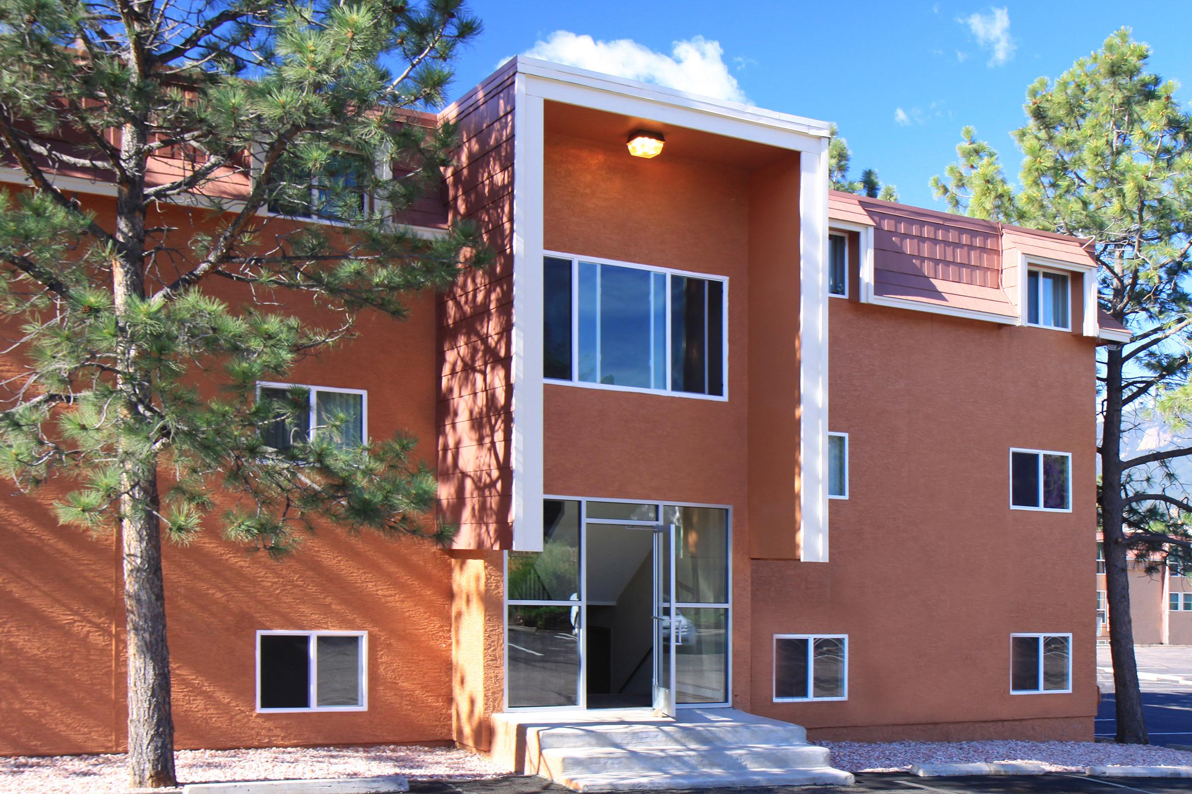 A modern, orange stucco building with large windows and a glass entrance door. Flanked by pine trees, the structure has a contemporary design with a flat roof. The setting is sunny with a clear blue sky. The entrance features steps leading up to the door, creating an inviting appearance.