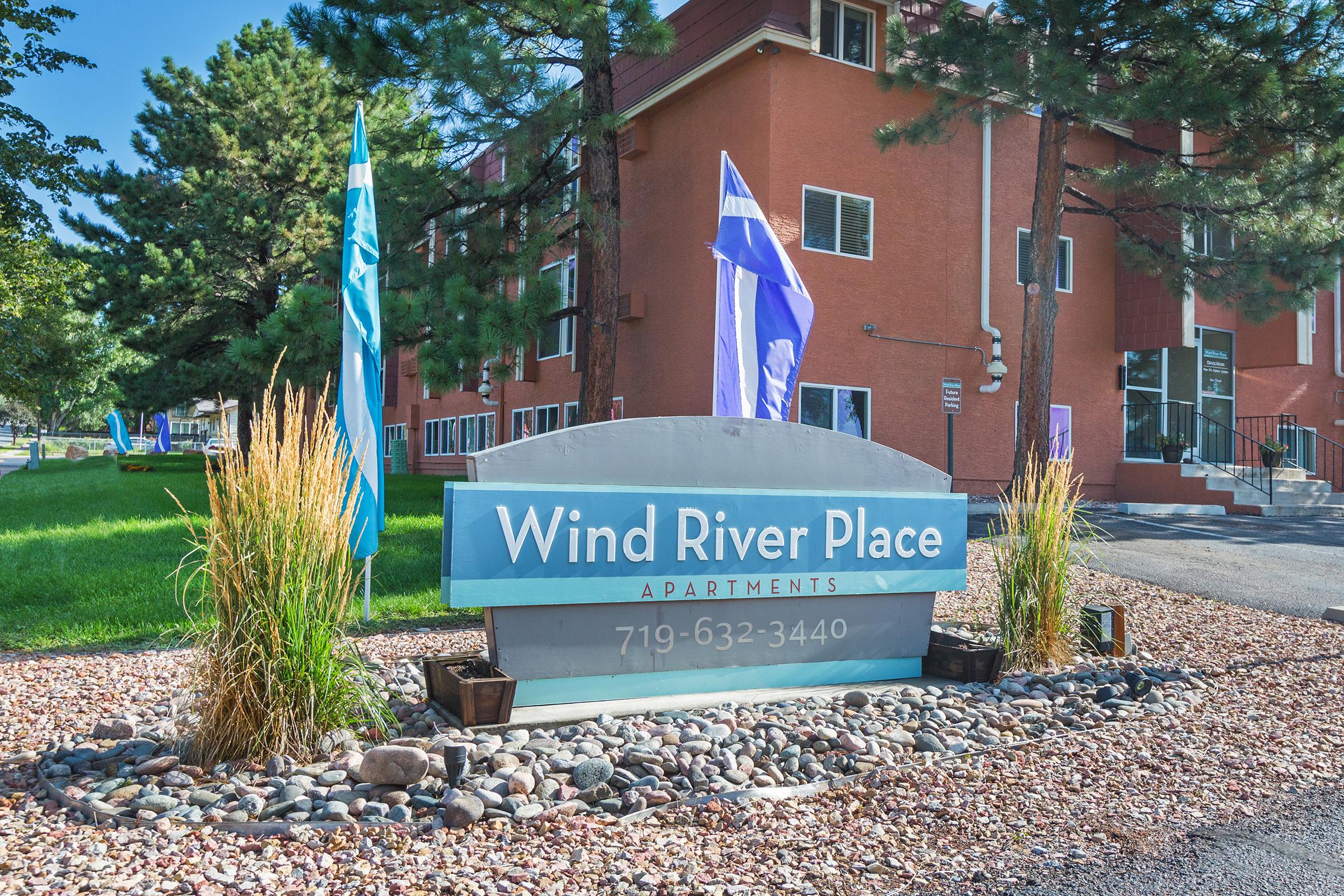 Sign for Wind River Place Apartments, displaying the name and contact number. The entrance features decorative plants, rocky landscaping, and colorful flags. The building is in the background, highlighting a well-maintained residential area.