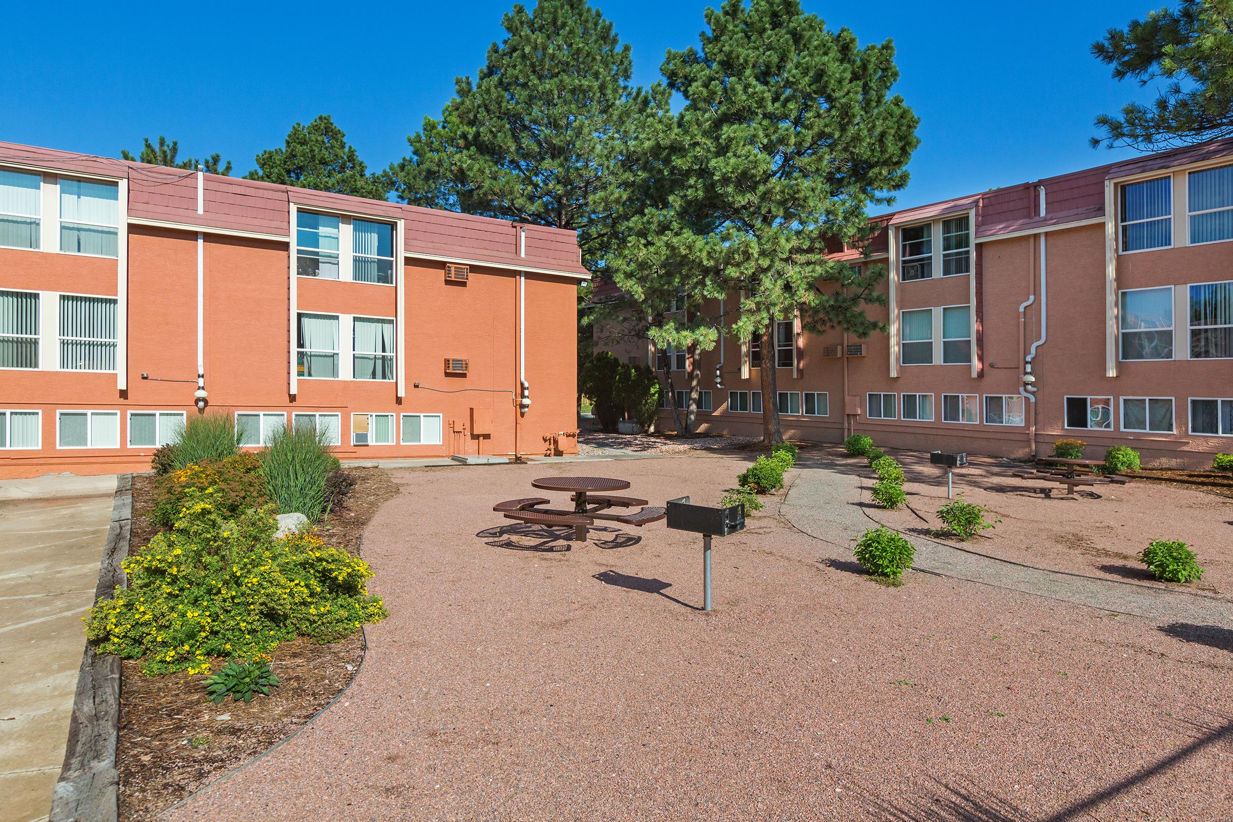 A view of a landscaped courtyard surrounded by two orange-colored buildings. The area features picnic tables, grill stations, and neatly arranged shrubs and flowers, with tall trees in the background under a clear blue sky.