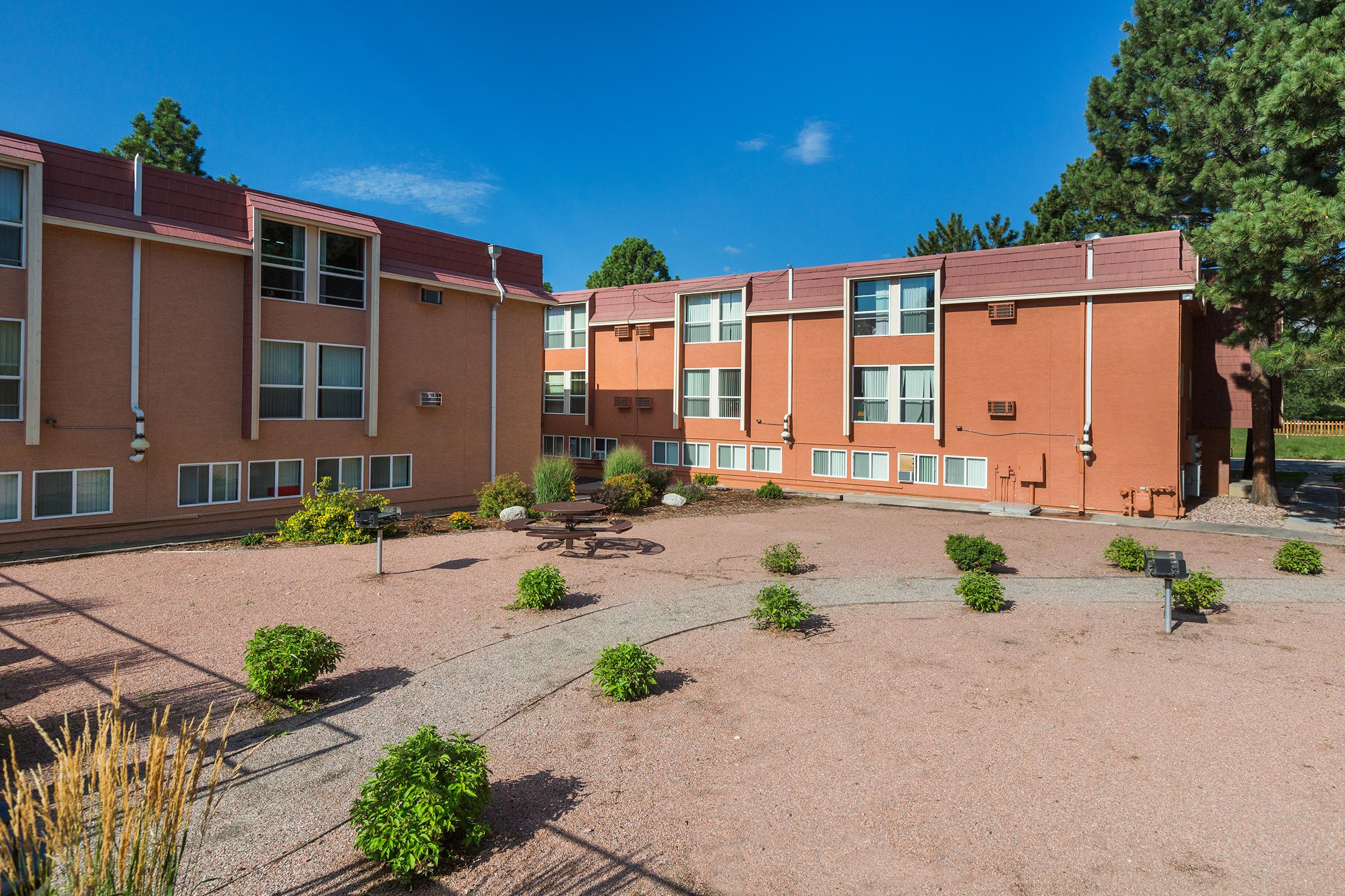 A landscaped courtyard surrounded by a two-story motel building. The area features small shrubs, gravel paths, and a few outdoor furniture pieces. The sky is clear and blue, enhancing the overall inviting atmosphere of the property.