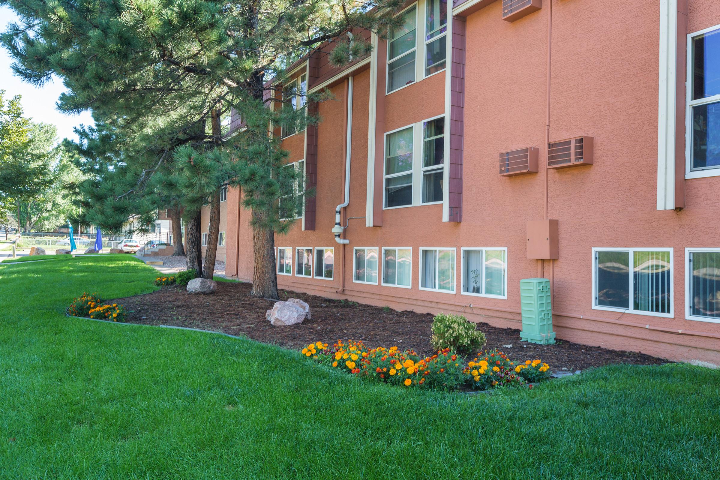 A well-maintained exterior of a building featuring a landscaped area. There are green lawns, flower beds with vibrant orange marigolds, and several trees lining the wall. The building has several windows, and there are air conditioning units installed beside them.