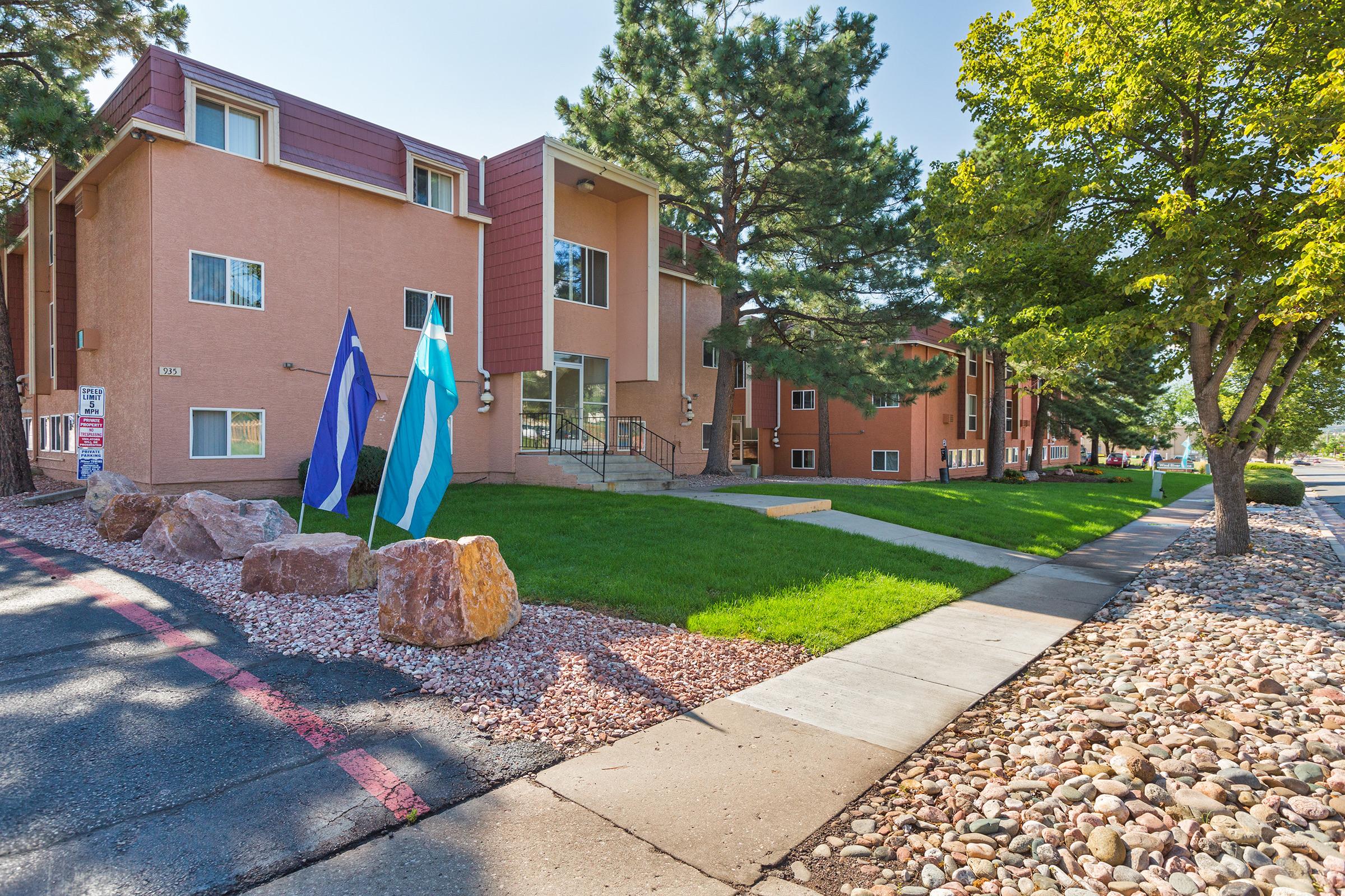 Exterior view of a modern apartment building featuring a landscaped lawn, trees, and decorative rocks. Flags in blue and teal are displayed in front of the entrance, which has steps leading up to it. Walkways are paved, and the overall setting appears well-maintained and inviting.