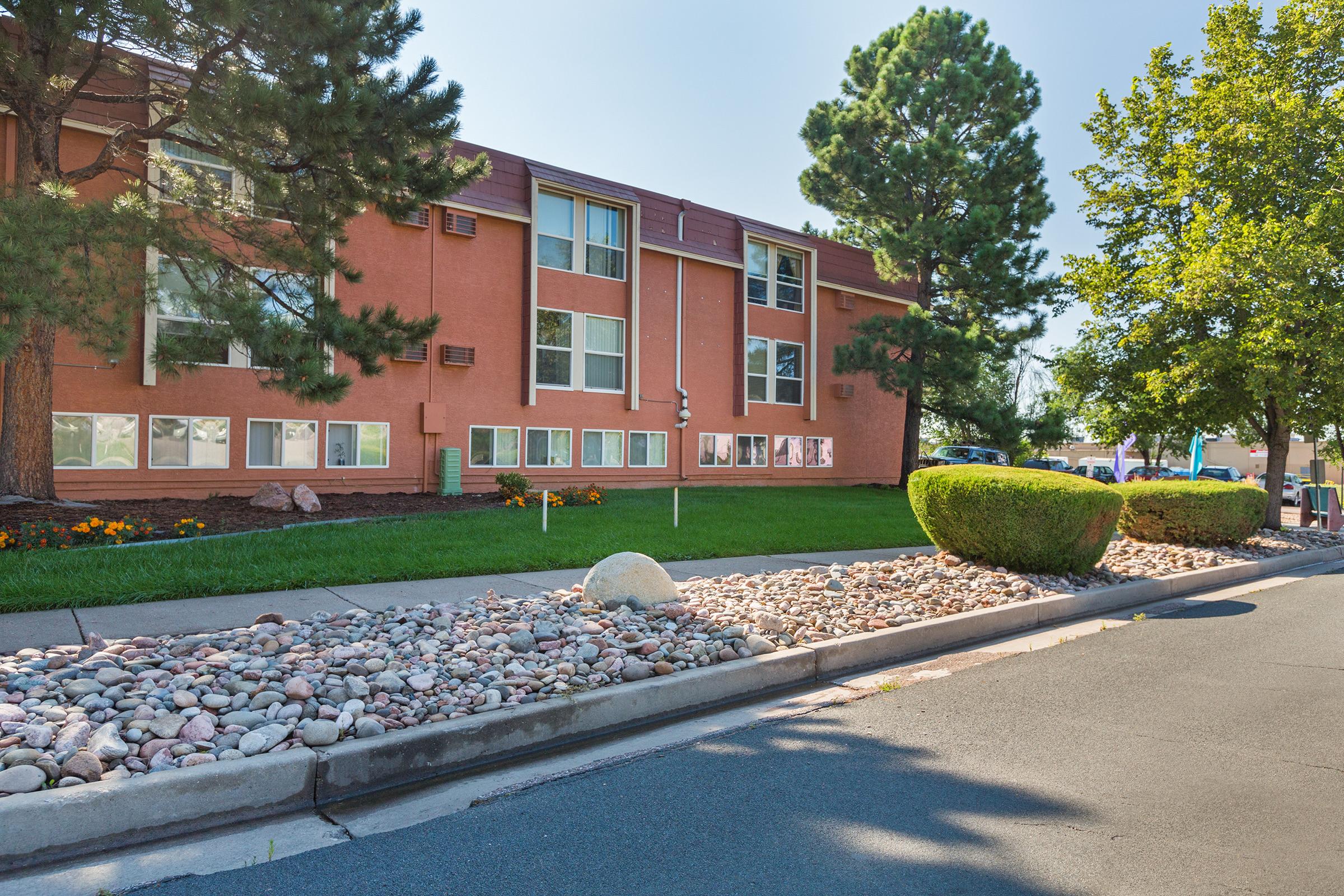 A view of a red-brick building with multiple windows, surrounded by green grass and shrubs. There is a landscaped area with rocks in front of the building, and trees provide shade. The scene is bright and sunny, indicating a pleasant day.