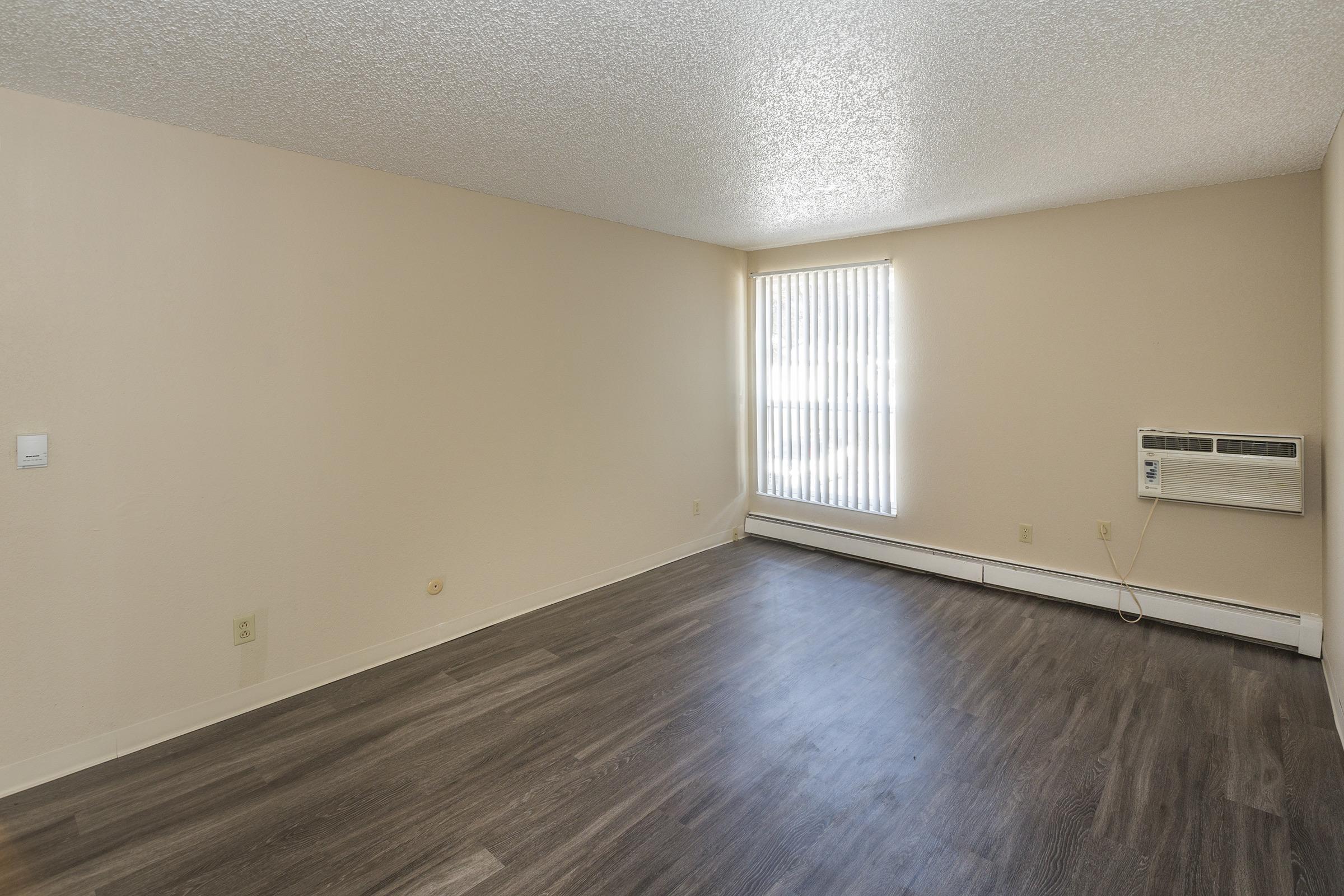 A spacious room with light-colored walls and a large window that allows natural light. The floor is covered with dark laminate flooring, and there is an air conditioning unit on one wall. The space appears empty and ready for furnishing.