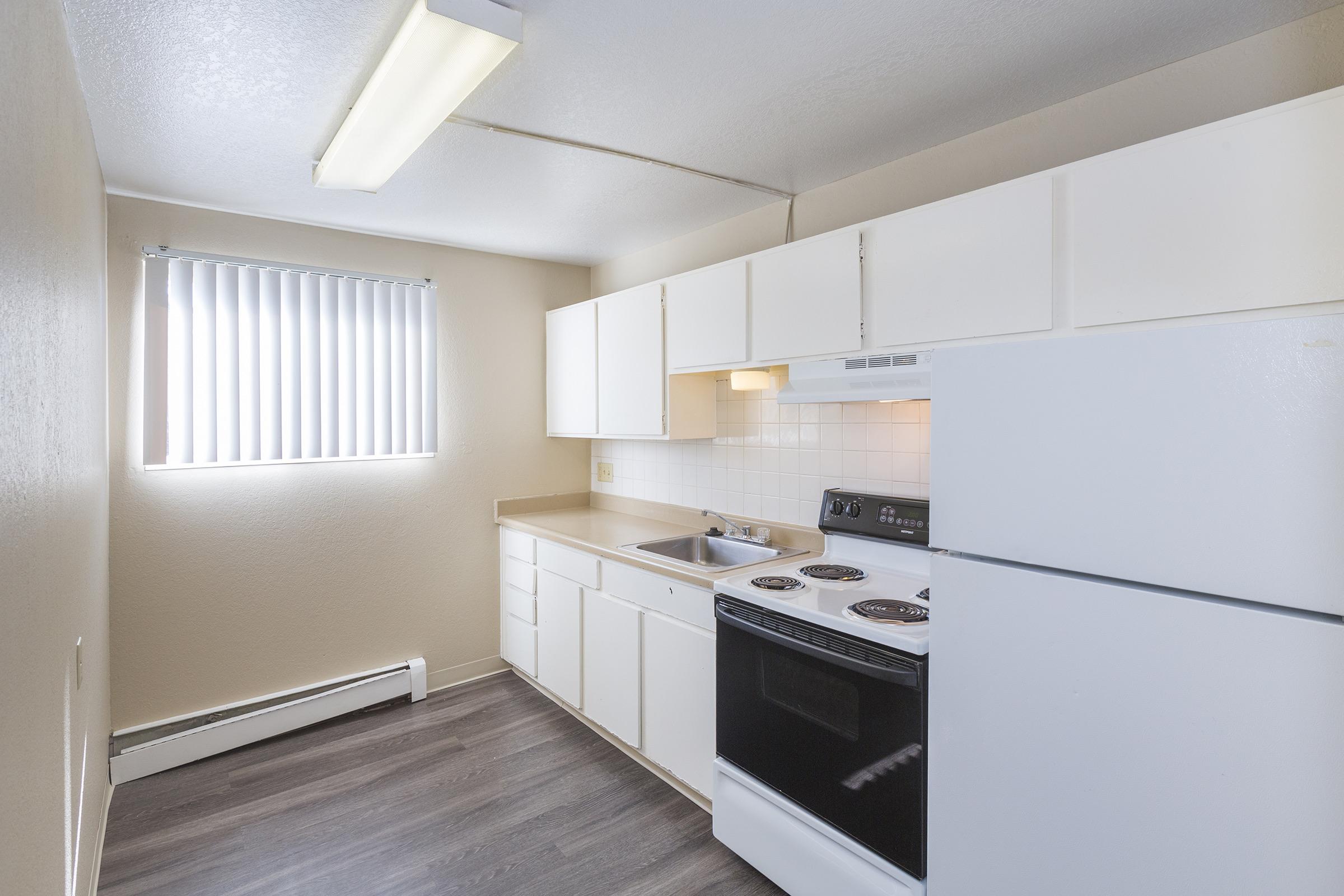A modern kitchen featuring white cabinets, a black stove and oven, a stainless steel sink, and a white refrigerator. The room has a neutral color scheme with light walls and a window with vertical blinds, allowing natural light to fill the space.