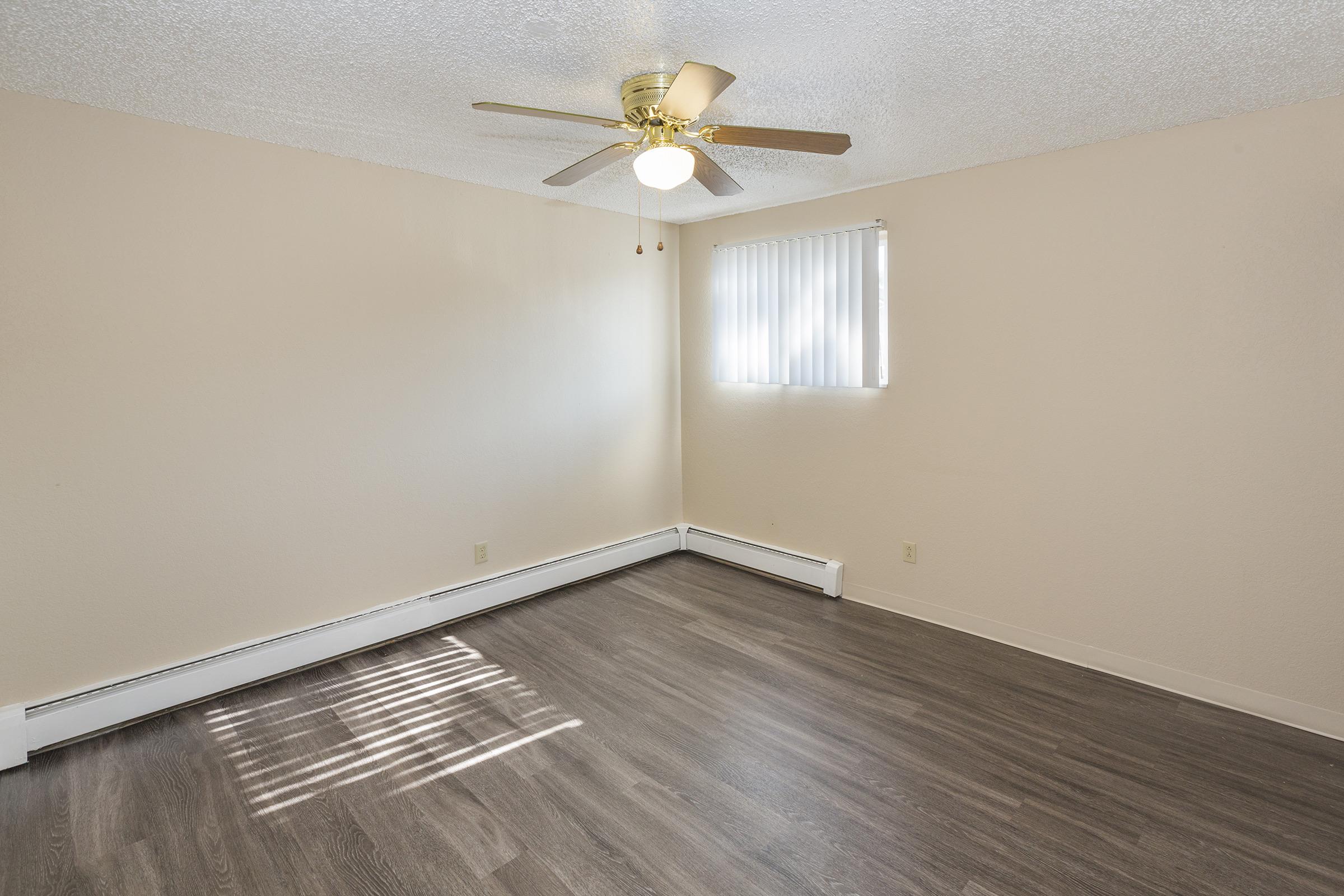 Empty room featuring light-colored walls and a ceiling fan. The floor is covered with dark laminate wood. A window on one wall allows natural light to filter in, casting shadows on the floor. No furniture is present, creating a spacious and airy atmosphere.