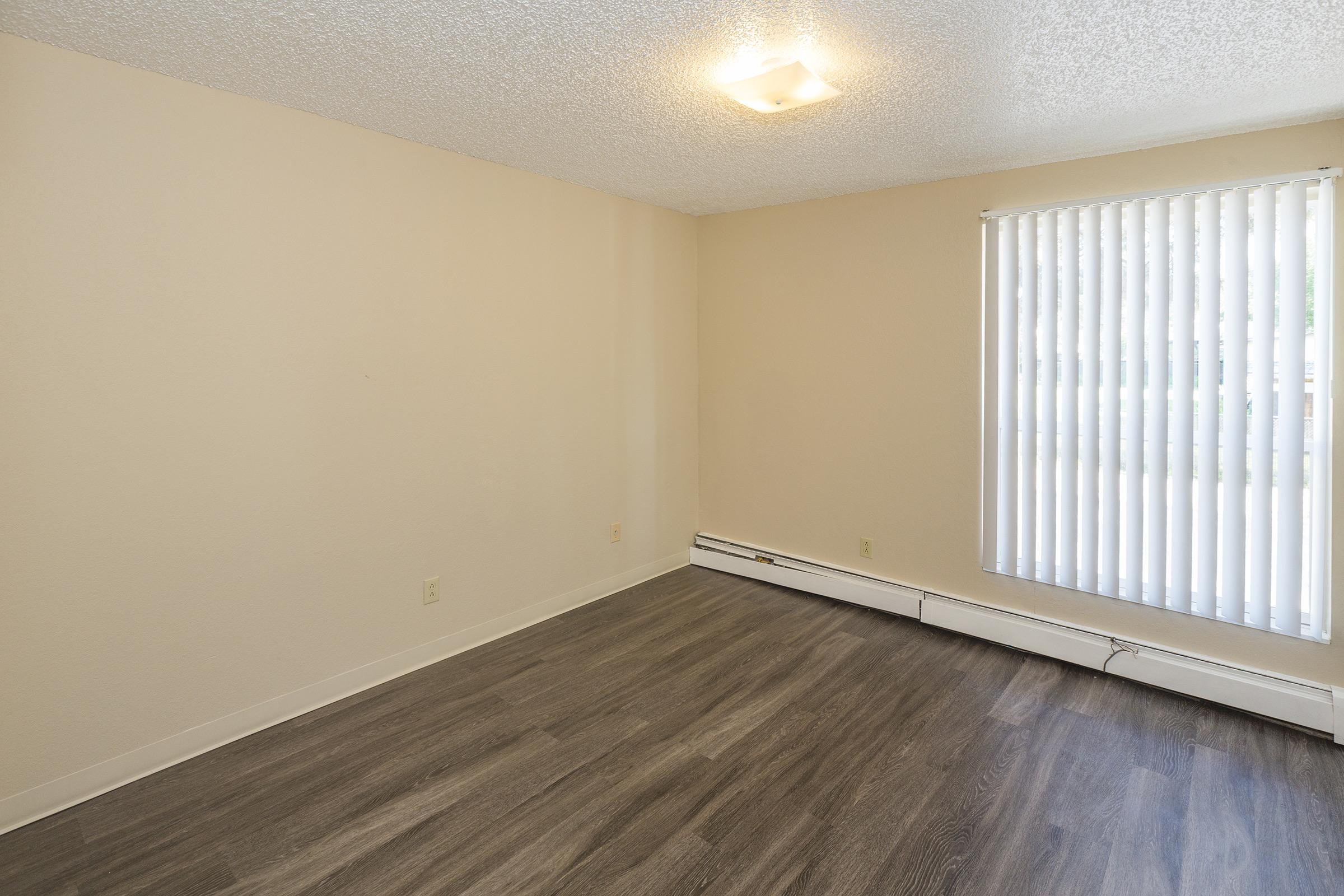 Empty room with light-colored walls and wooden-style flooring. A window with vertical blinds lets in natural light. The ceiling features a small light fixture, and there are no furnishings present, emphasizing the spaciousness of the area.