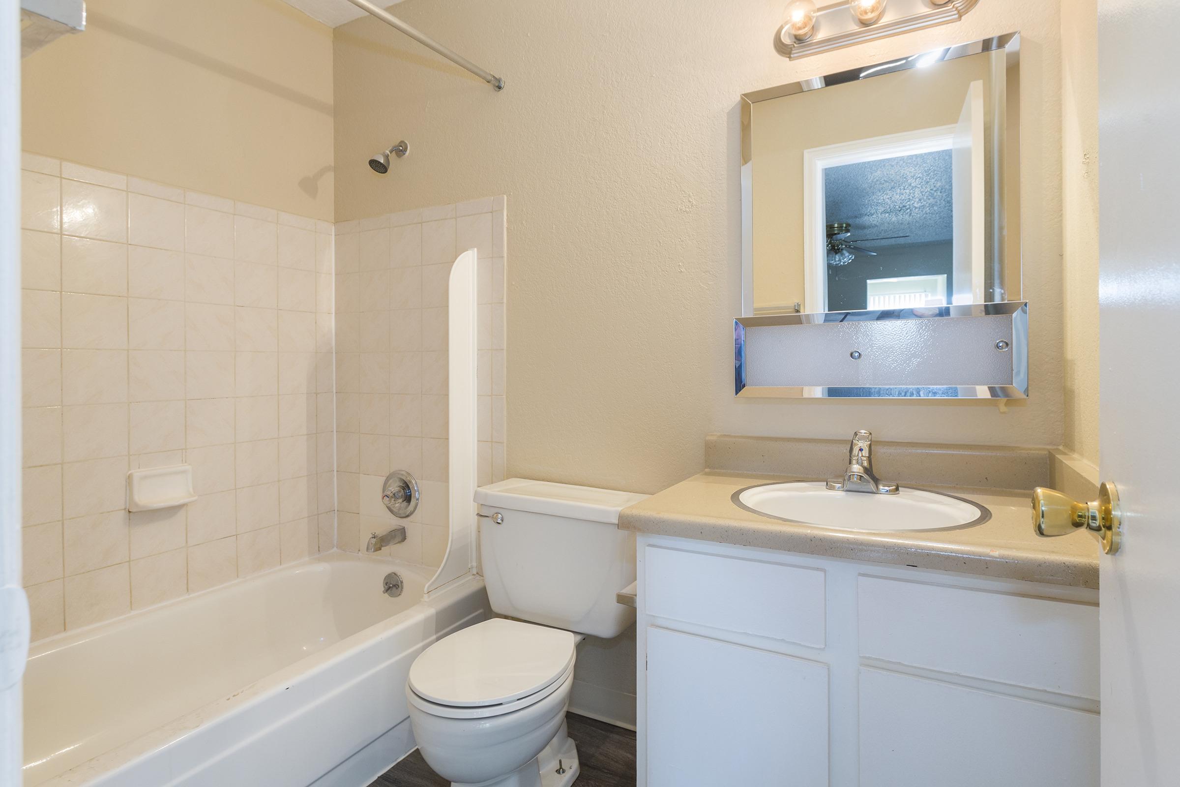A clean and simple bathroom featuring a bathtub with a shower, a toilet, and a sink with a countertop, all in light-colored tiles and walls. A mirror is mounted above the sink, and there is a small window in the background. The floor has a wooden appearance, adding warmth to the space.