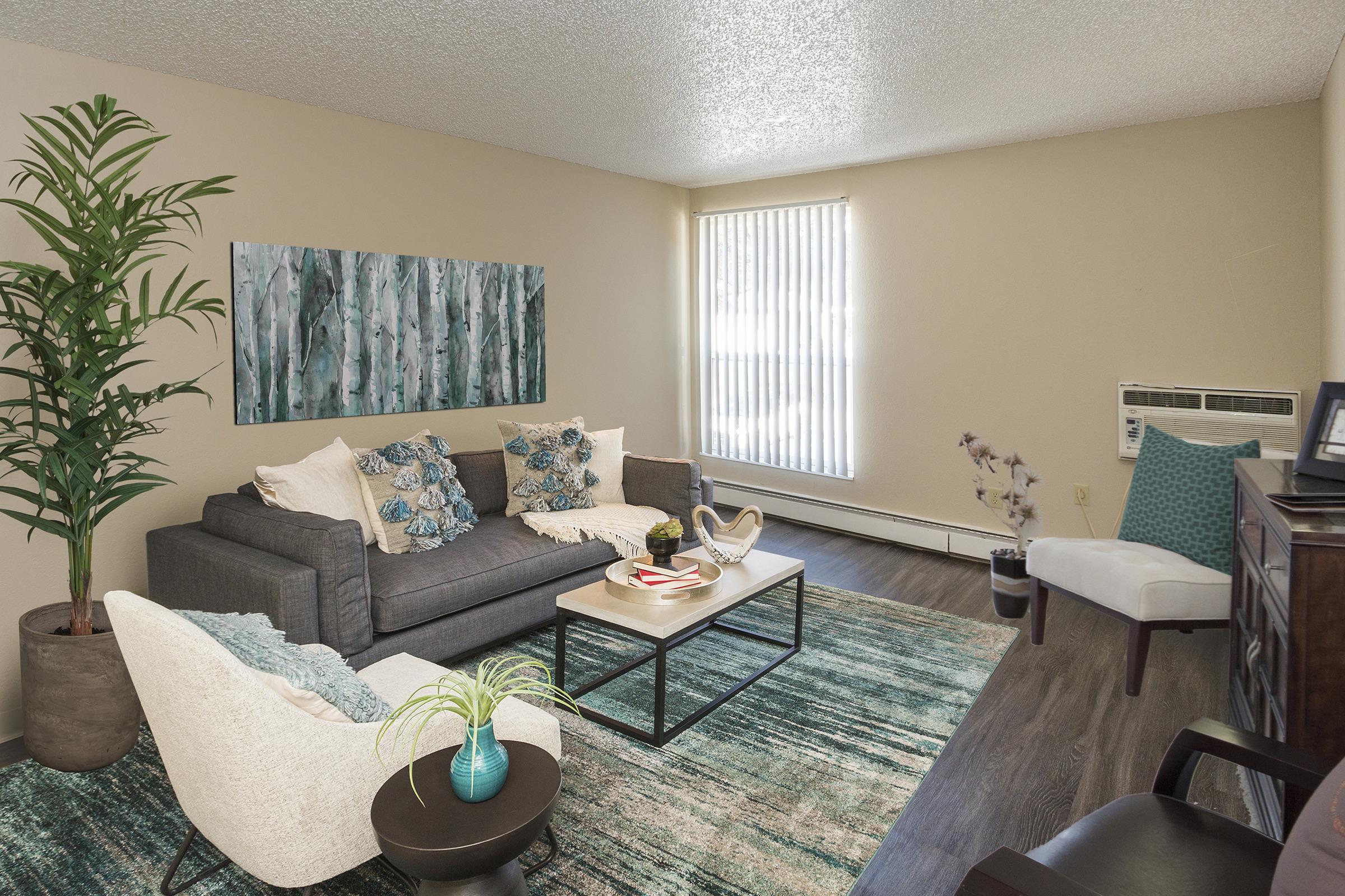 A cozy living room featuring a gray sofa with decorative blue and white pillows, a coffee table with books and decor items, a light-colored armchair, and a potted plant. The walls are a neutral tone, with a piece of artwork depicting abstract trees, and a large window allows natural light to enter the space.