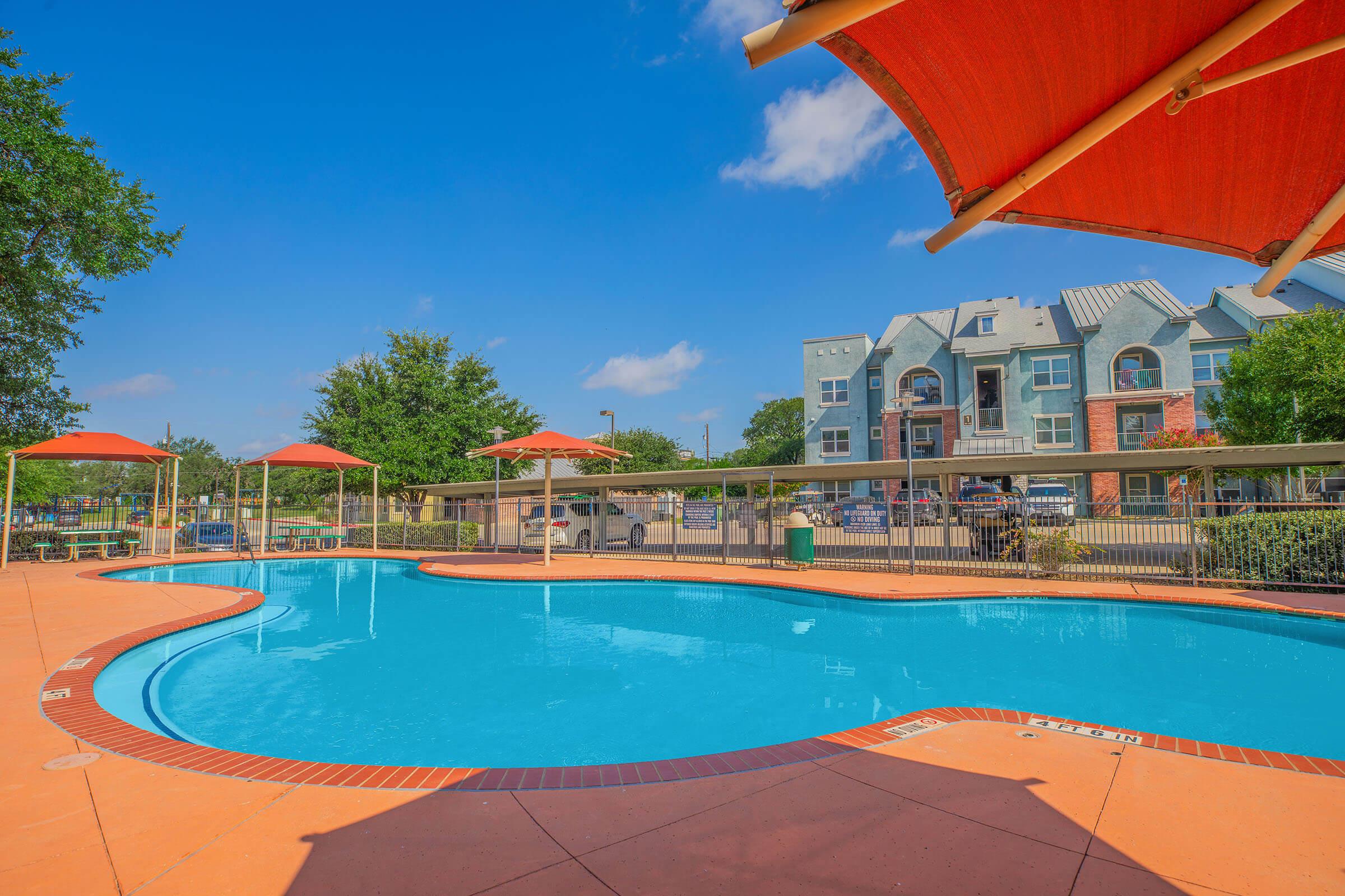A sparkling blue swimming pool surrounded by orange umbrellas and lounge chairs, with a vibrant outdoor area. In the background, modern multi-story buildings and green trees are visible under a clear blue sky.