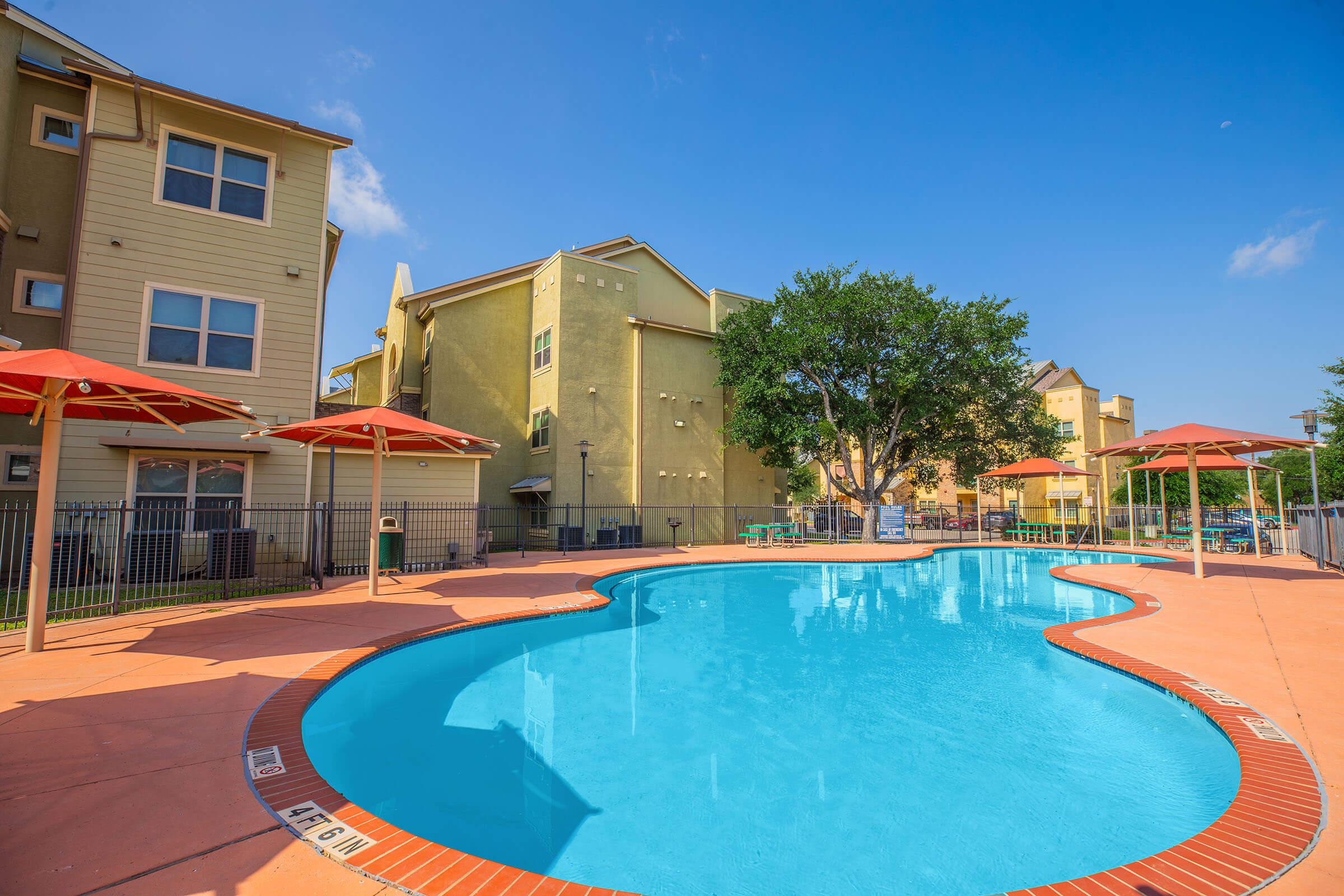 A bright and inviting outdoor swimming pool surrounded by a landscaped area. The pool features a textured, curved edge and is bordered by colorful lounge chairs and umbrellas. Nearby, there are modern apartment buildings under a clear blue sky, creating a relaxed and vibrant atmosphere.