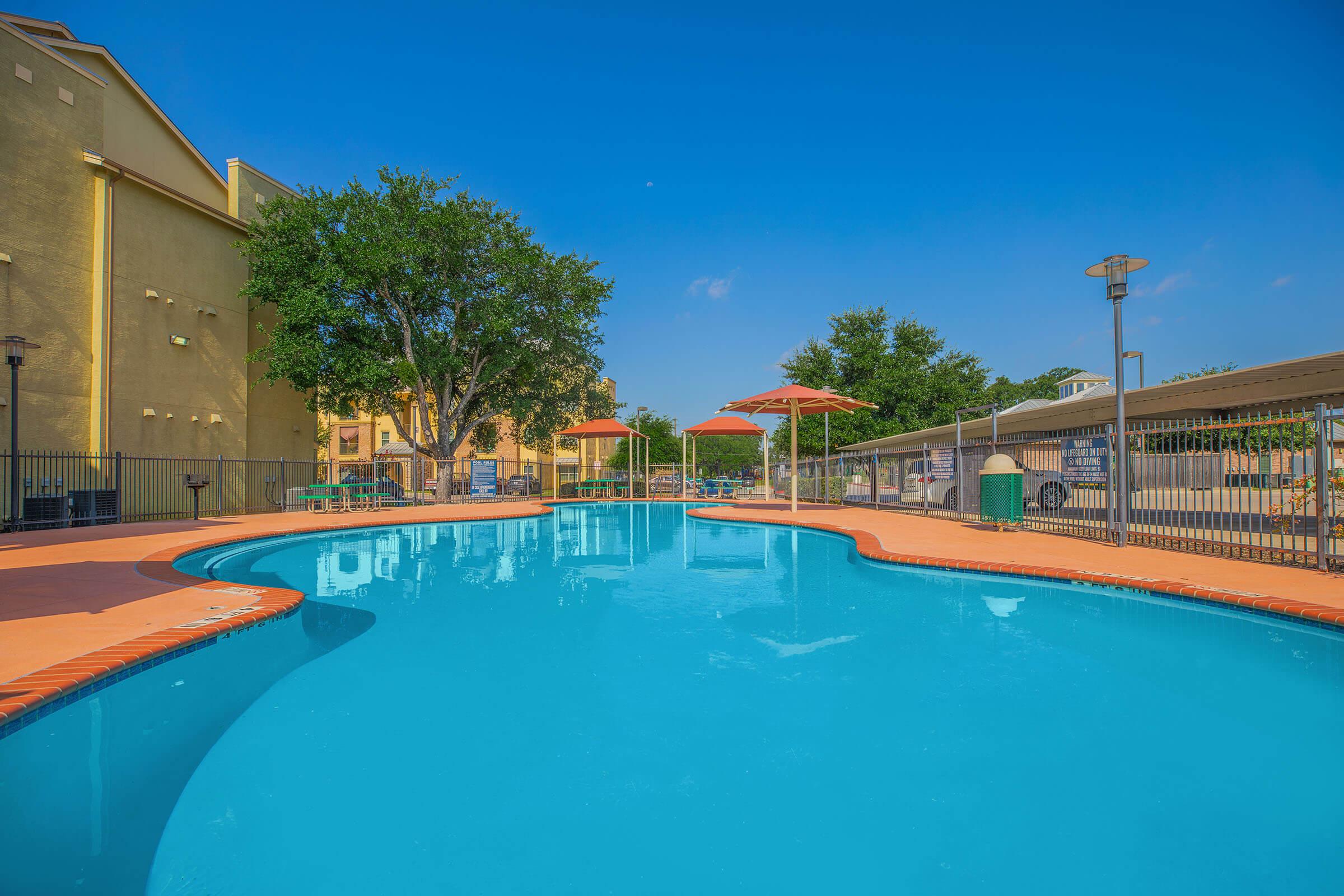 A vibrant swimming pool surrounded by a well-maintained area, featuring orange loungers and umbrellas. Lush green trees and a clear blue sky provide a serene atmosphere, while a tall building is visible in the background. The water is calm and inviting, perfect for relaxation or a refreshing swim.