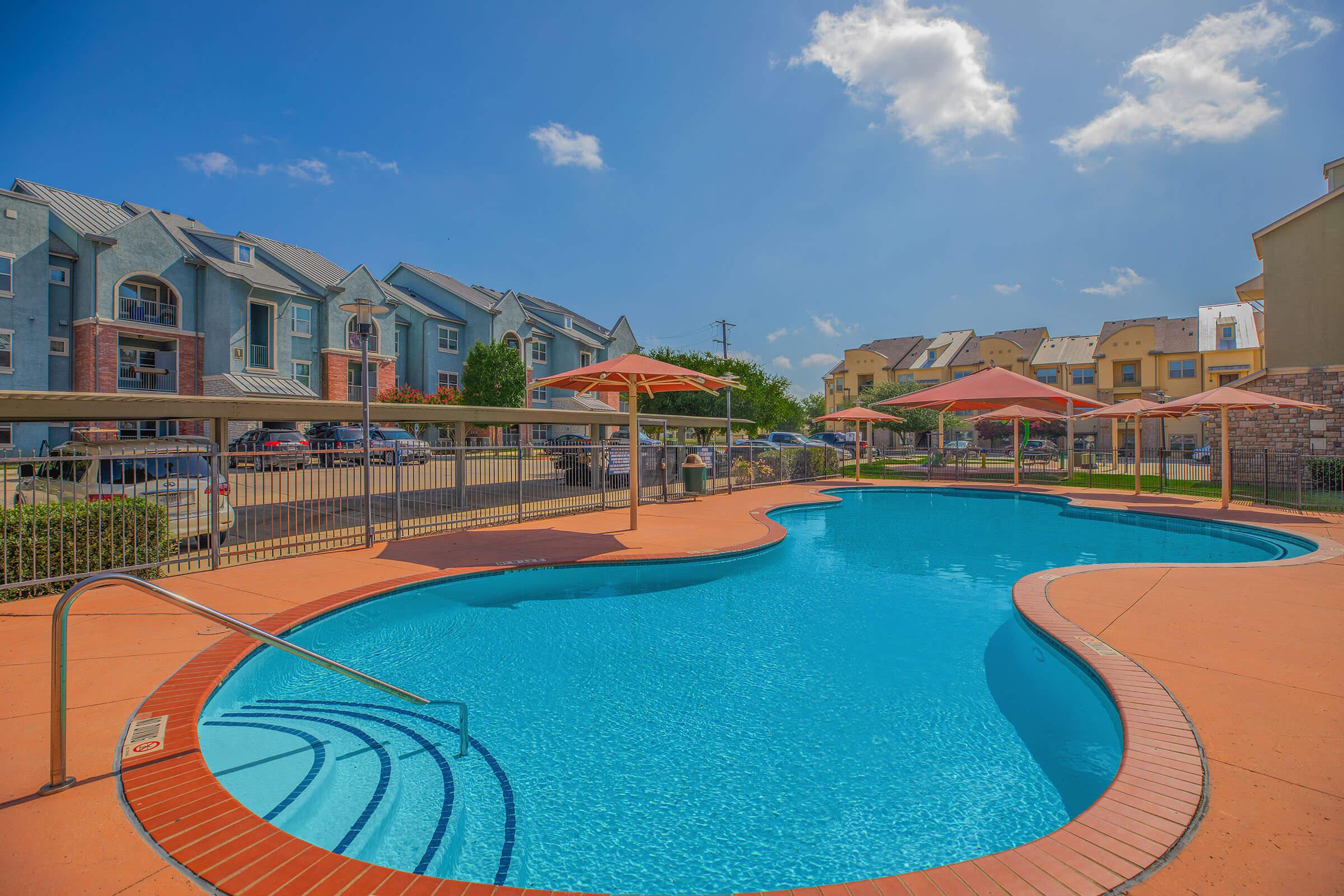 A clear blue swimming pool surrounded by orange umbrellas and lounge areas, with colorful residential buildings in the background. The sky is bright and sunny with a few clouds, creating a relaxing outdoor atmosphere.