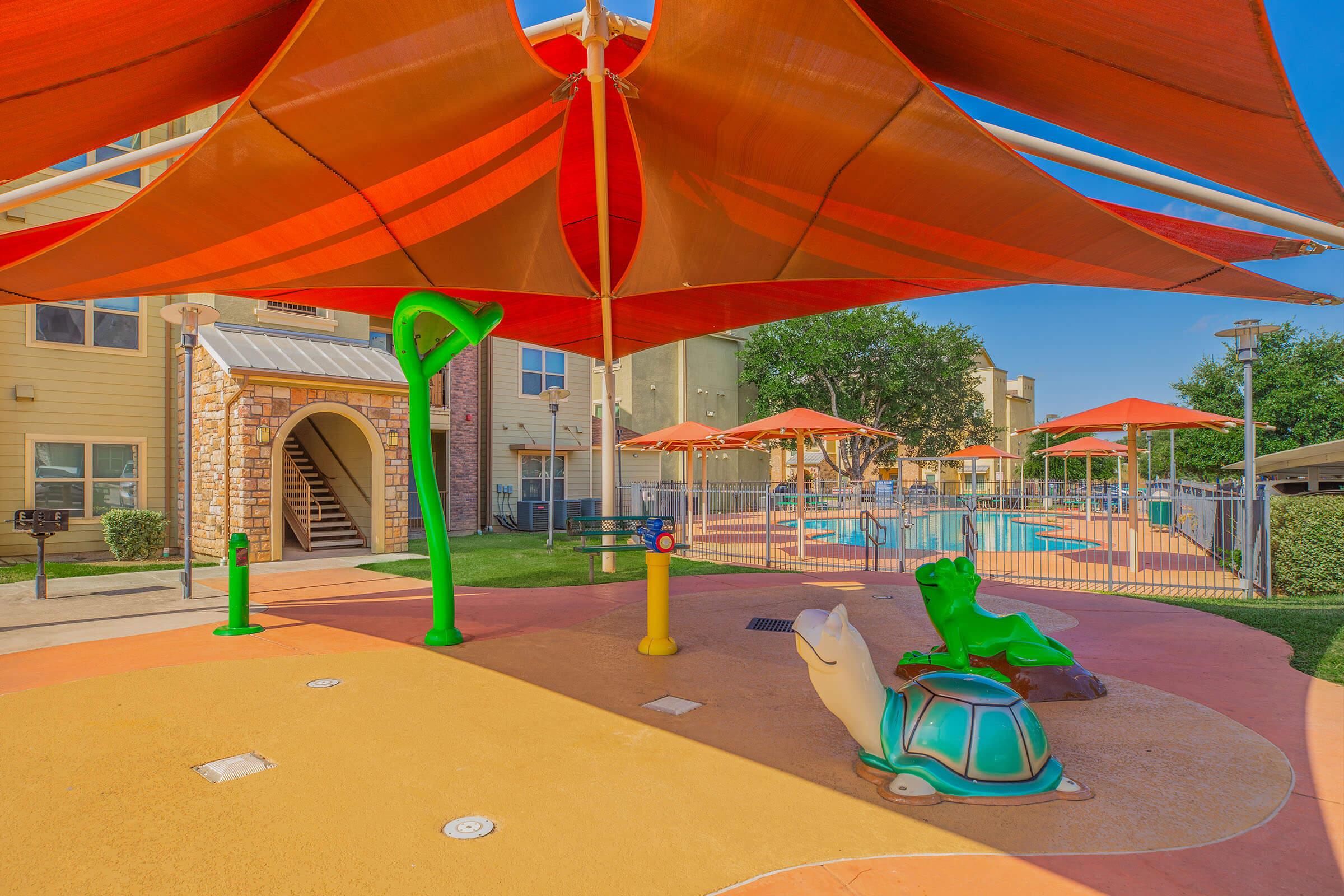Colorful playground with a turtle and frog sculpture, shaded by a large red canopy. In the background, there is a swimming pool surrounded by various orange umbrellas and apartment buildings. The area is vibrant and designed for children's play.