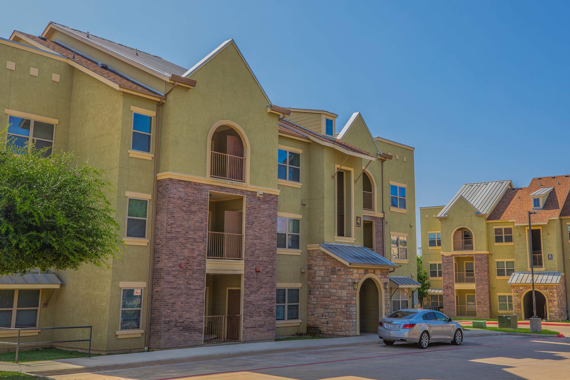 Modern multi-story apartment building with a combination of green and brick exteriors. The design features arch-shaped balconies and large windows, set against a clear blue sky. A silver car is parked in front of the building, surrounded by well-maintained landscaping.