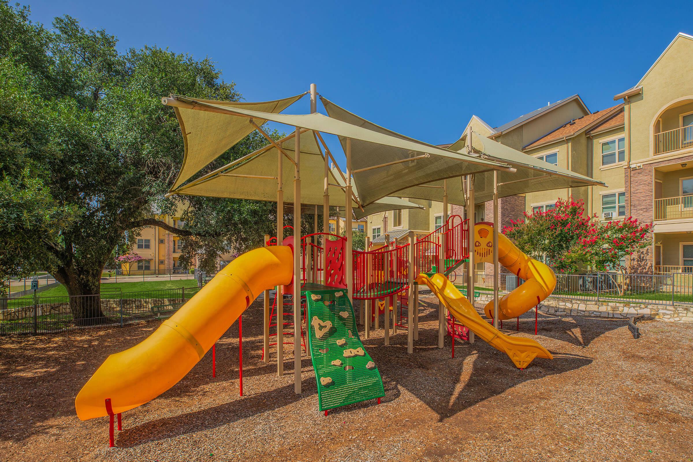 Playground with colorful slides and climbing structures under a large canopy. The slides are bright orange and yellow, surrounded by gravel and grass. Nearby, there are residential buildings and flowering trees, creating a cheerful outdoor space for children to play.