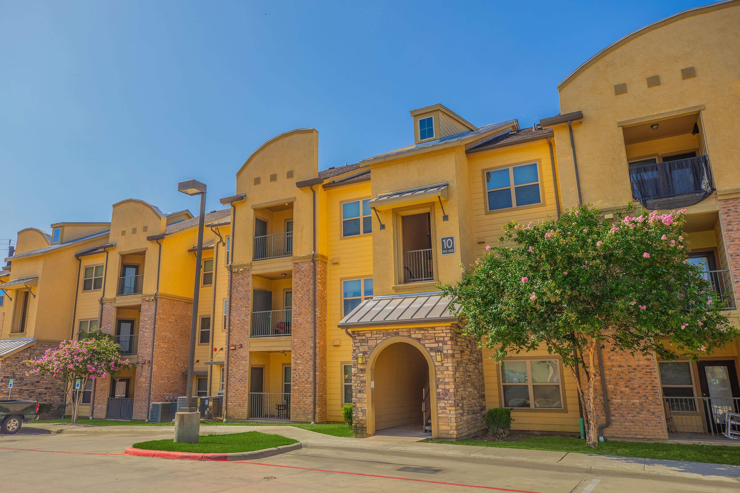 A brightly lit apartment complex featuring multiple stories with a mixture of brick and yellow siding. Balconies are visible, along with flowering trees in the landscape. The driveway leads to the entrance of the building, with clear blue skies above.