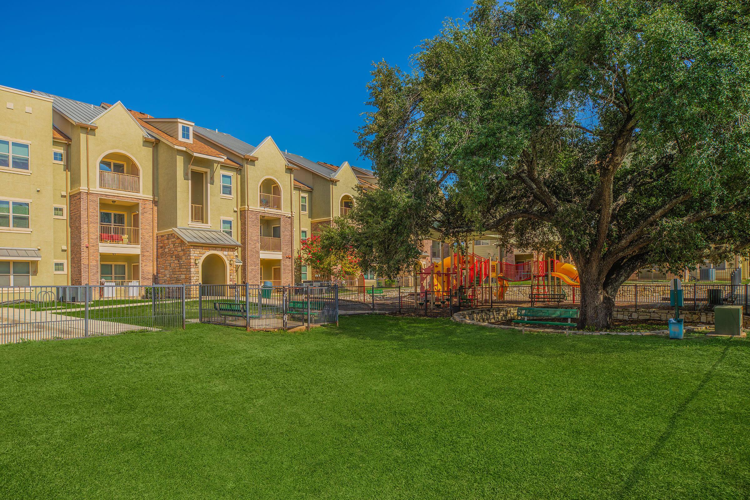A vibrant playground surrounded by green grass with a large tree nearby, set against a backdrop of colorful apartment buildings under a clear blue sky. The playground features slides and climbing structures, creating a lively outdoor space for children.