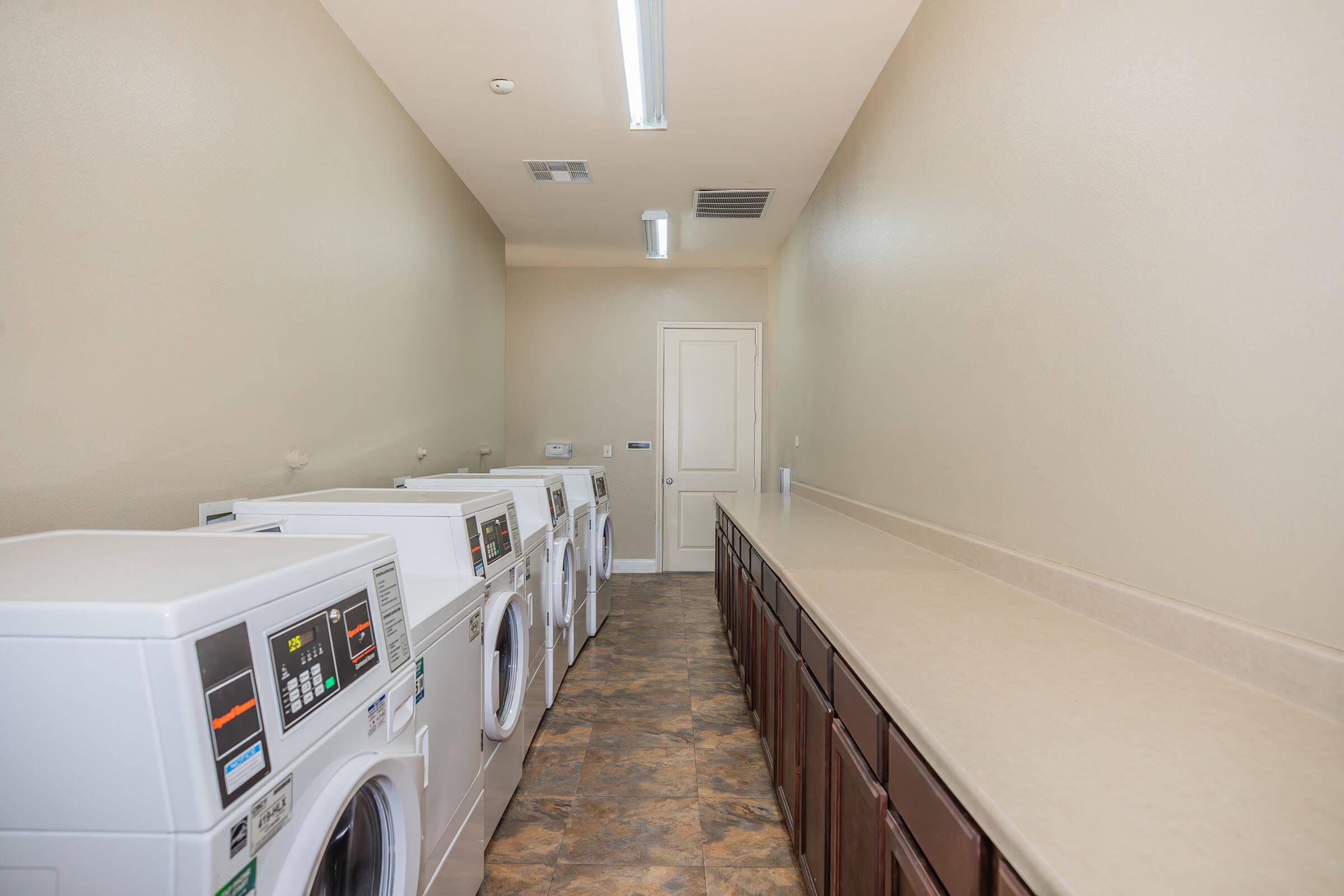 A clean, well-lit laundry room featuring several white washing machines and dryers aligned against the wall. There is a long countertop for folding clothes and wooden cabinets above. The walls are a light beige color, and the floor is tiled in a darker shade.
