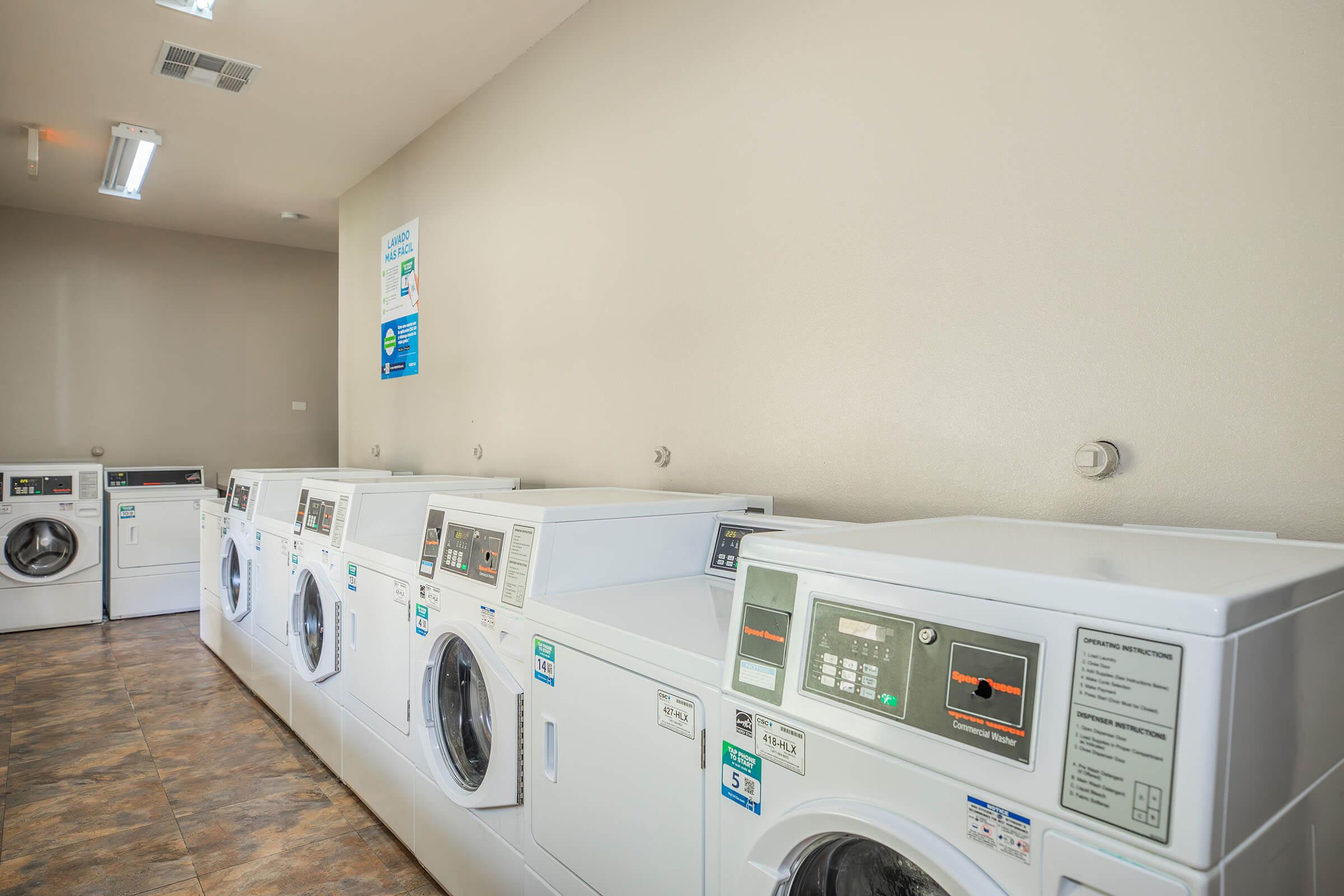 A row of modern washing machines against a neutral-colored wall in a laundry room. The machines are clean and well-maintained, featuring digital displays. The floor is tiled, and there is overhead lighting illuminating the space. A sign is visible on the wall, providing information or instructions related to the laundry service.