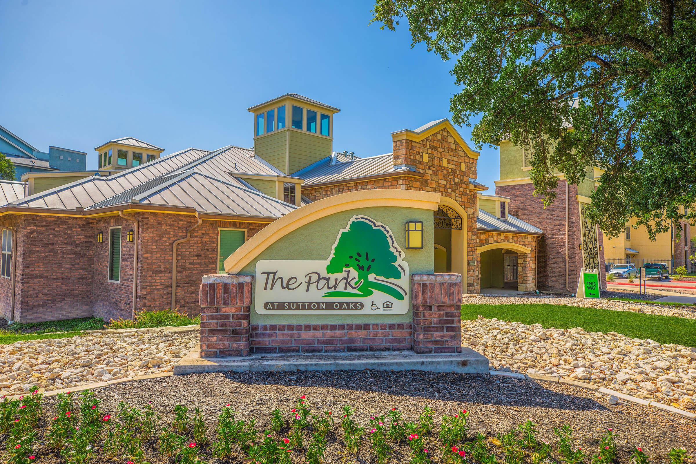 Sign for "The Park at Sutton Oaks" located in front of a stylish building with a modern design, featuring a landscaped area with rocks and flowers. Bright blue sky overhead and various architectural elements visible, including a tower.