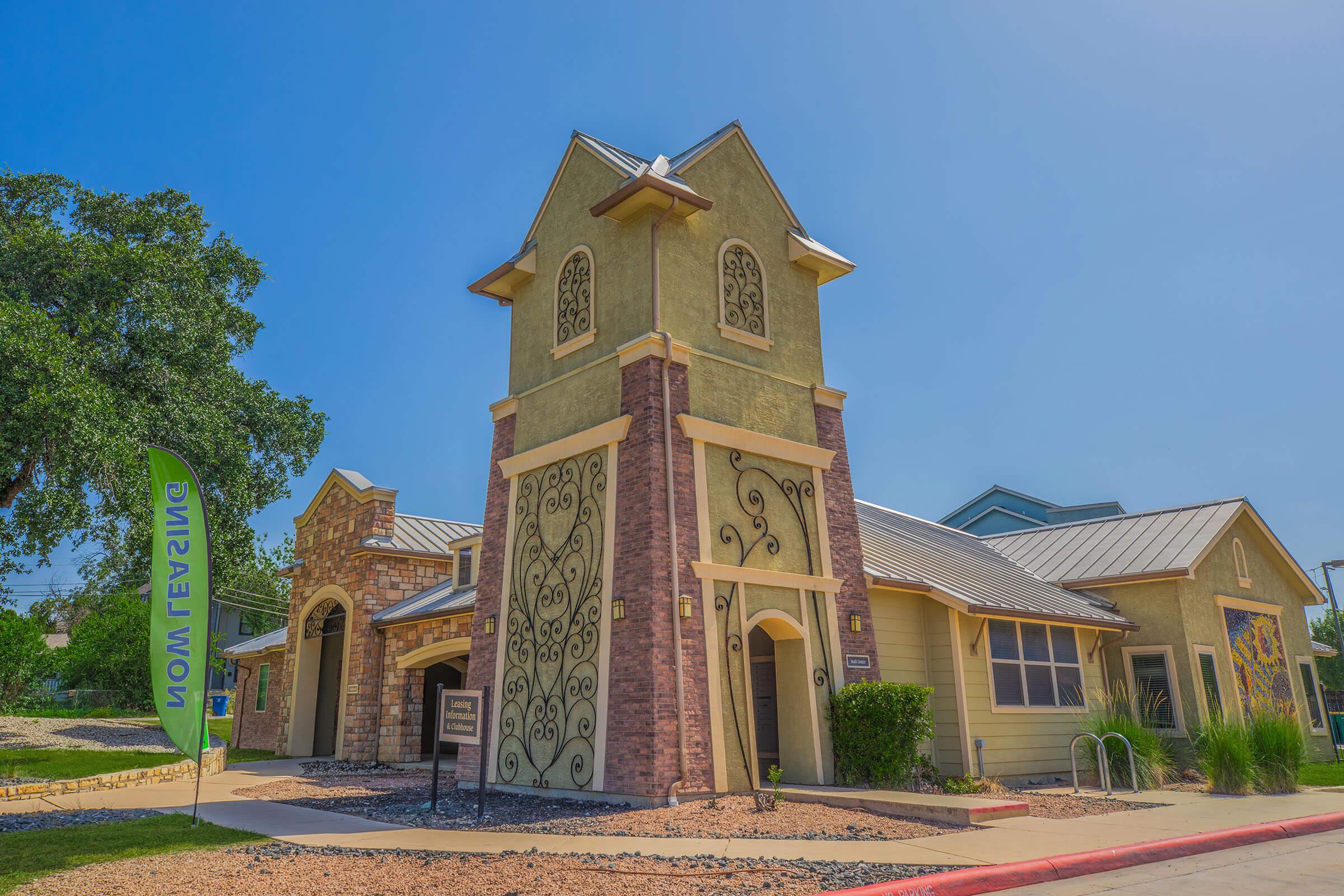 A colorful building with unique architectural features, including a tall tower and decorative elements on the walls. The structure is surrounded by greenery and has a signpost nearby. A flag is visible in the foreground, adding to the vibrant atmosphere of the scene.