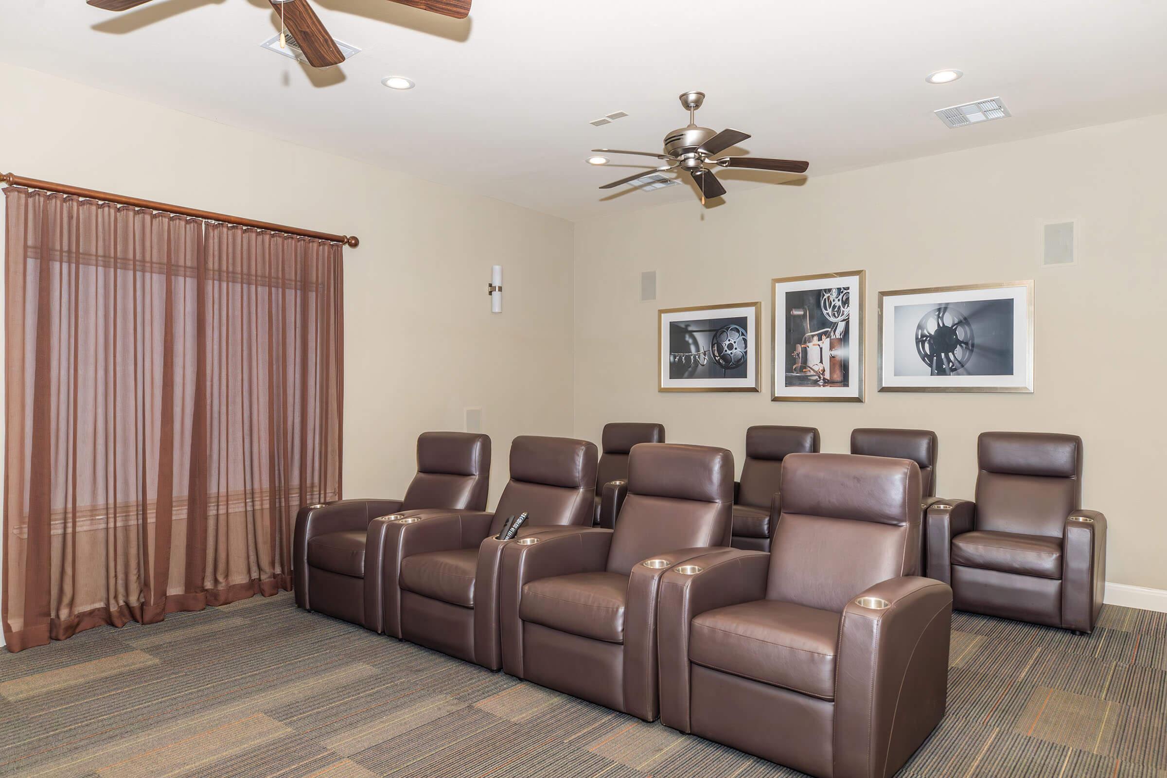 A cozy home theater room featuring brown leather recliners arranged in a row. The room has a ceiling fan and soft lighting, with dark curtains covering a window. There are framed black-and-white photographs on the walls, enhancing the elegant atmosphere of the space. The flooring is carpeted in neutral tones.