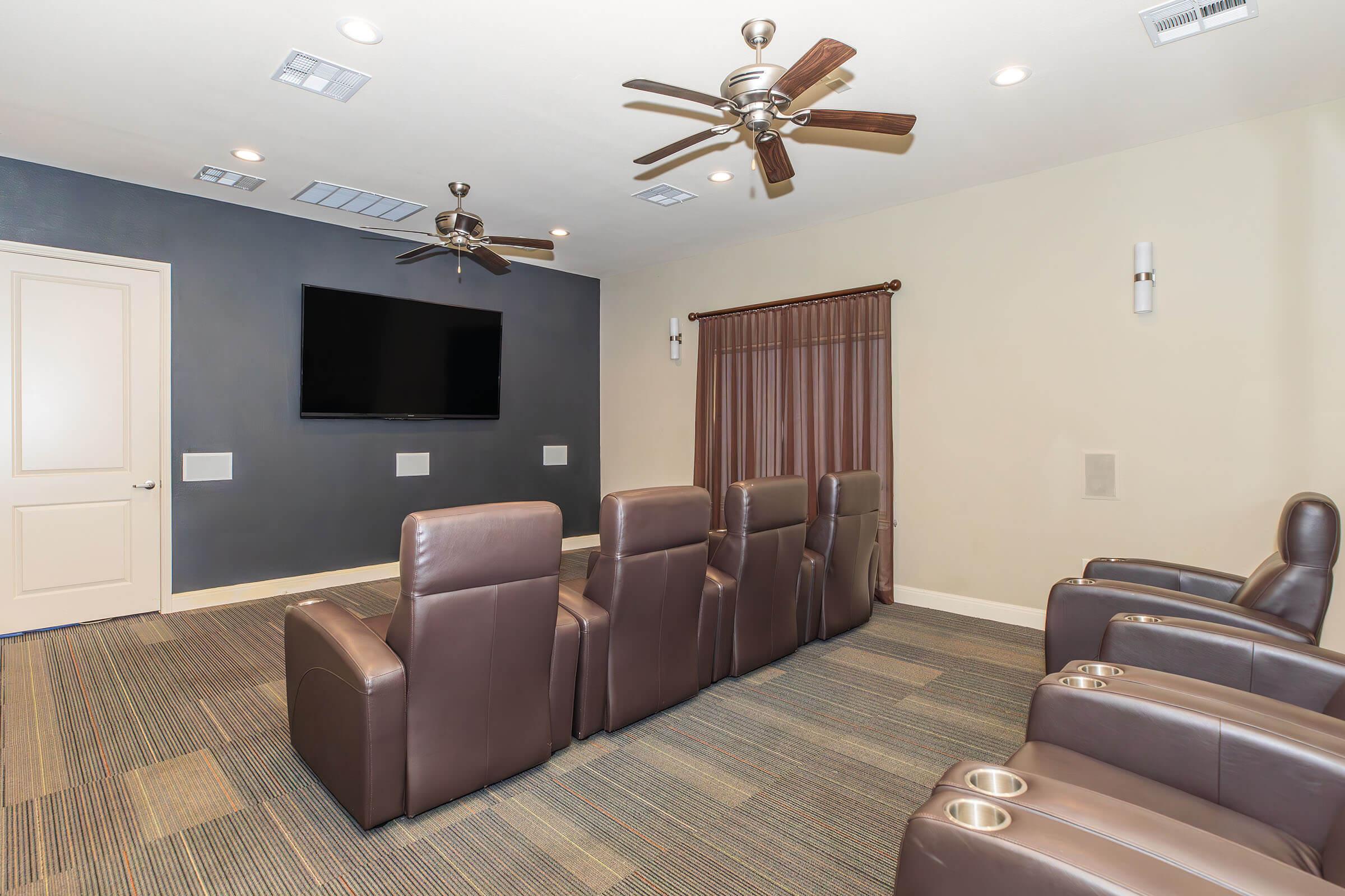 Home theater room featuring plush brown reclining chairs arranged in front of a flat-screen TV mounted on the wall. The room has light-colored walls, ceiling fans, and a curtain on one side, with carpeting in a muted pattern. Soft lighting adds a cozy atmosphere.