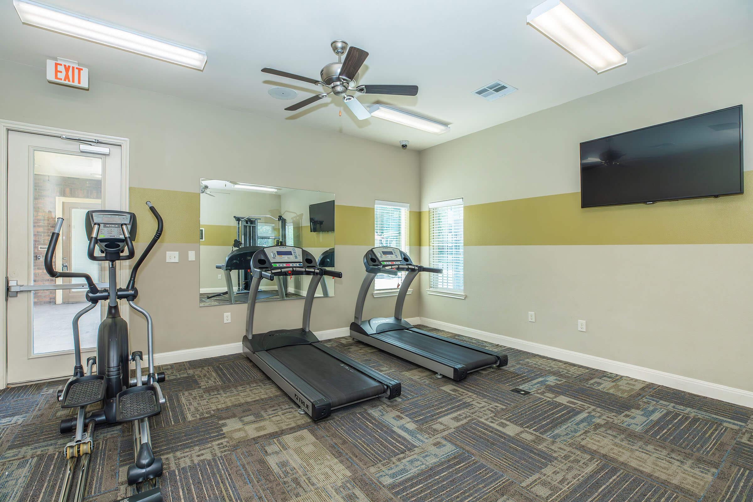 A well-lit fitness room featuring two treadmills, an elliptical machine, a large wall-mounted television, a ceiling fan, and mirrors. The walls are painted in neutral colors with a yellow accent, and the floor has a patterned carpet. Large windows allow natural light into the space.
