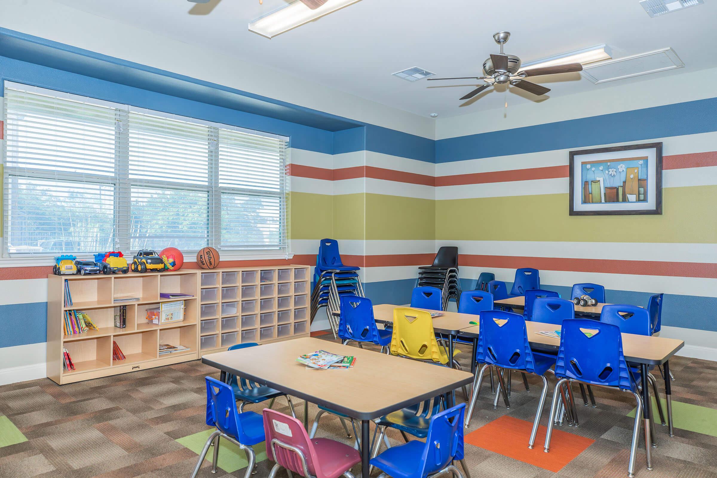 A brightly colored classroom featuring blue and green striped walls. There are small tables with yellow and blue chairs arranged for students. A bookshelf holds toys and books, and additional chairs are stacked nearby. Large windows let in natural light, creating a welcoming learning environment.