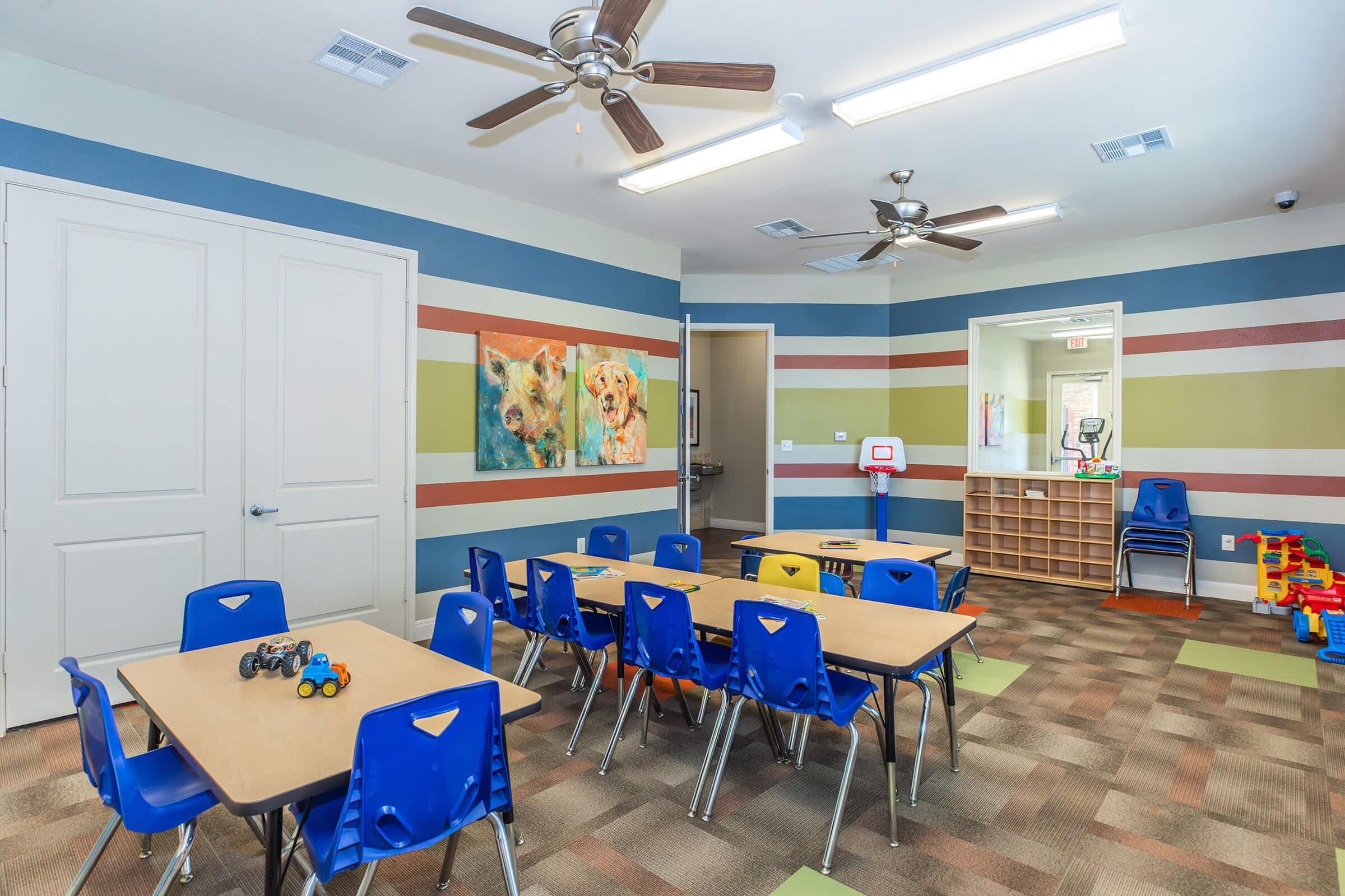 Brightly colored classroom featuring several small tables and blue chairs. The walls are decorated with horizontal stripes in various colors and two framed animal pictures. In the background, there is a doorway leading to another room, a small basketball hoop, and shelves with toys. Ceiling fans are visible.