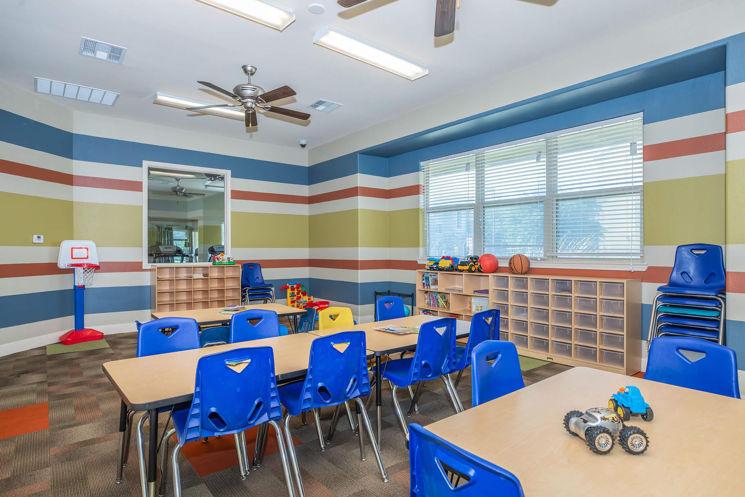 Playroom with colorful striped walls, featuring a basketball hoop, several toy bins, and a spacious layout. There are blue chairs and tables arranged for activities, along with various toys visible on a table. A large window allows natural light to fill the room, creating a bright and inviting atmosphere.