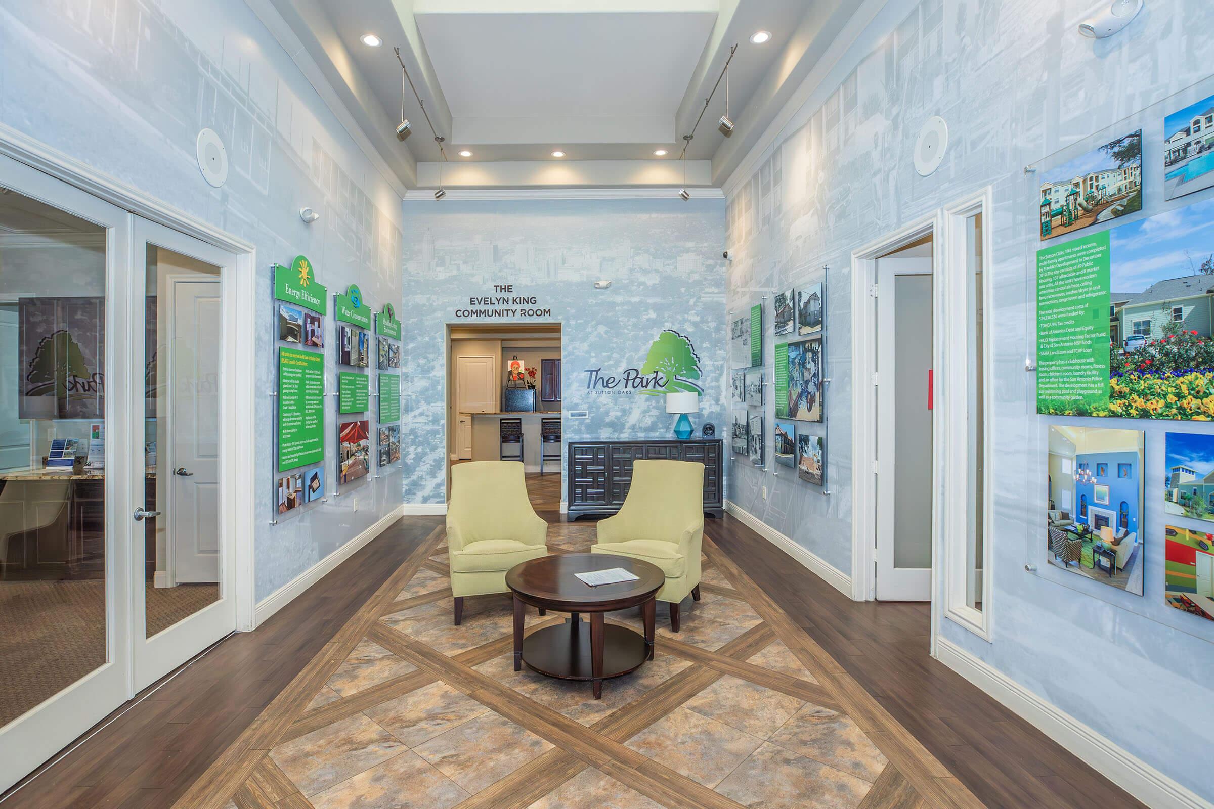 A well-lit reception area with two yellow chairs facing a circular table. The walls are decorated with light-colored murals and informational panels featuring photos and text. A sign above a door indicates "The Everling Company Zoom," and there are details about the local community displayed. The floor has a decorative pattern.