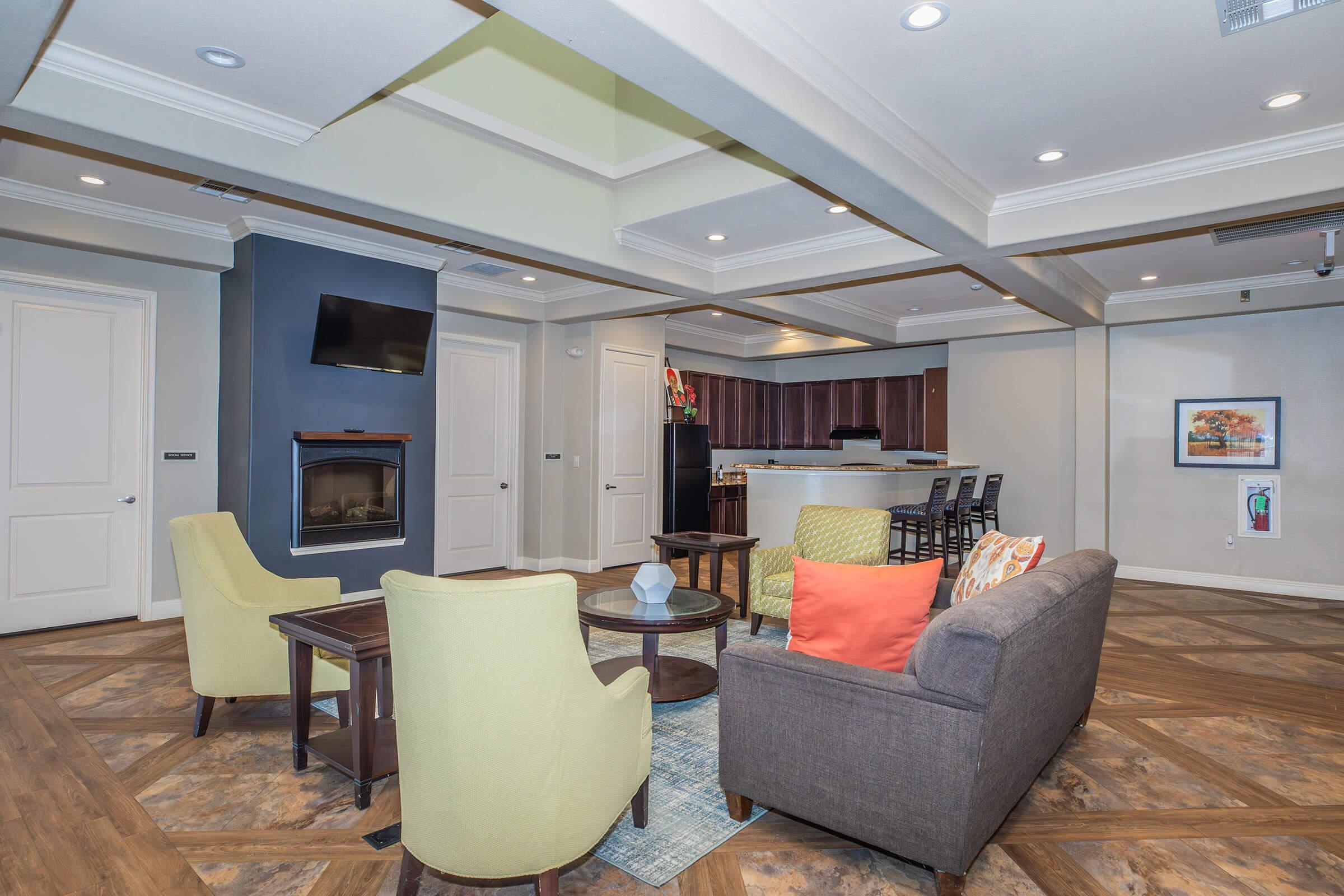 A cozy lounge area featuring a gray sofa, two green armchairs, and a wooden coffee table. There’s a light brown fireplace, a flat-screen TV mounted on the wall, and a kitchen bar with high stools in the background. Warm lighting and decorative artwork enhance the inviting atmosphere.