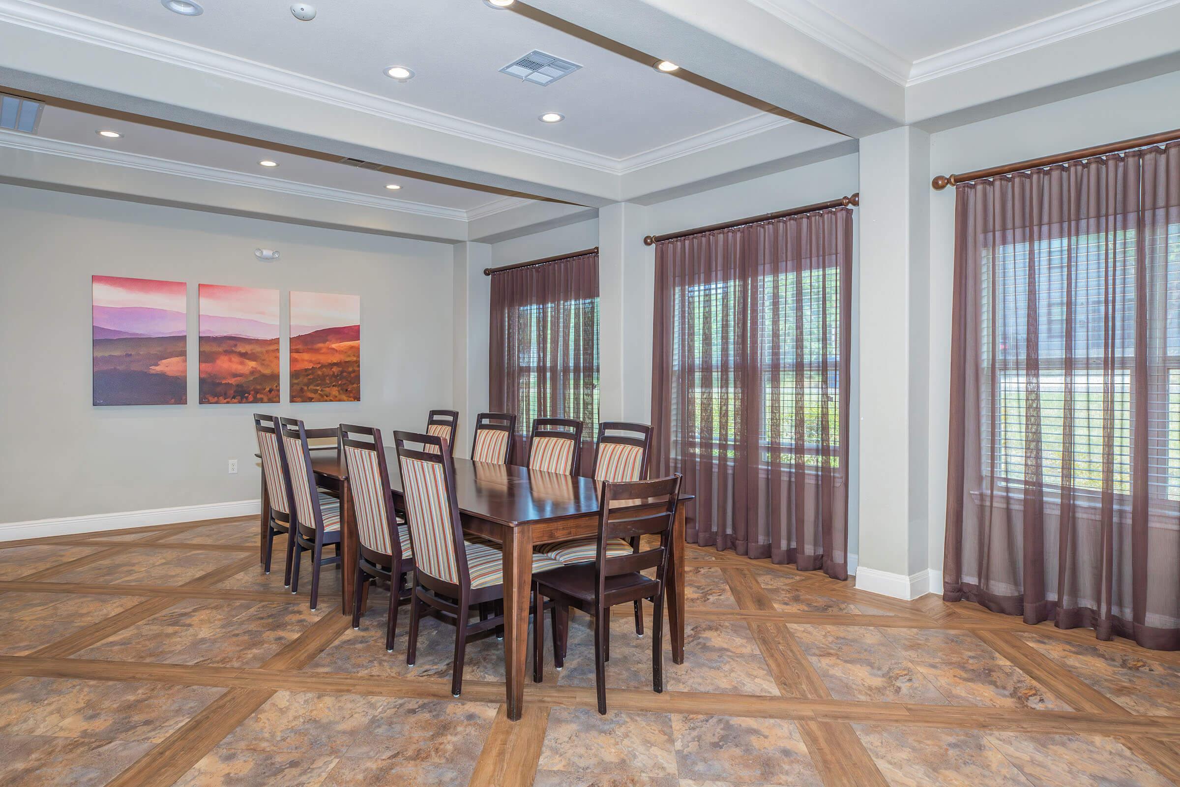 A modern dining room featuring a large wooden table surrounded by ten striped chairs. The walls are painted a soft gray, adorned with two large abstract landscape paintings. Natural light streams in through floor-length sheer curtains, enhancing the inviting atmosphere of the space.