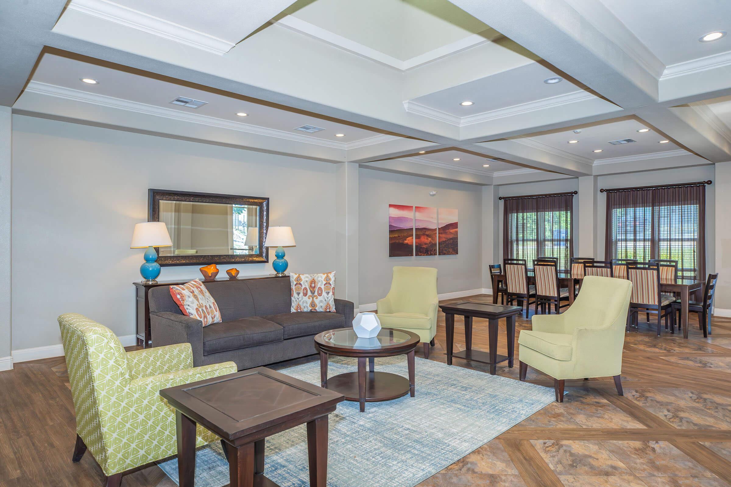 A modern living area featuring a gray sofa with colorful pillows, two yellow armchairs, a round coffee table, and a decorative rug. There are stylish lamps on side tables, and in the background, a dining area with a large table and chairs, along with artwork on the walls and natural light coming from the windows.
