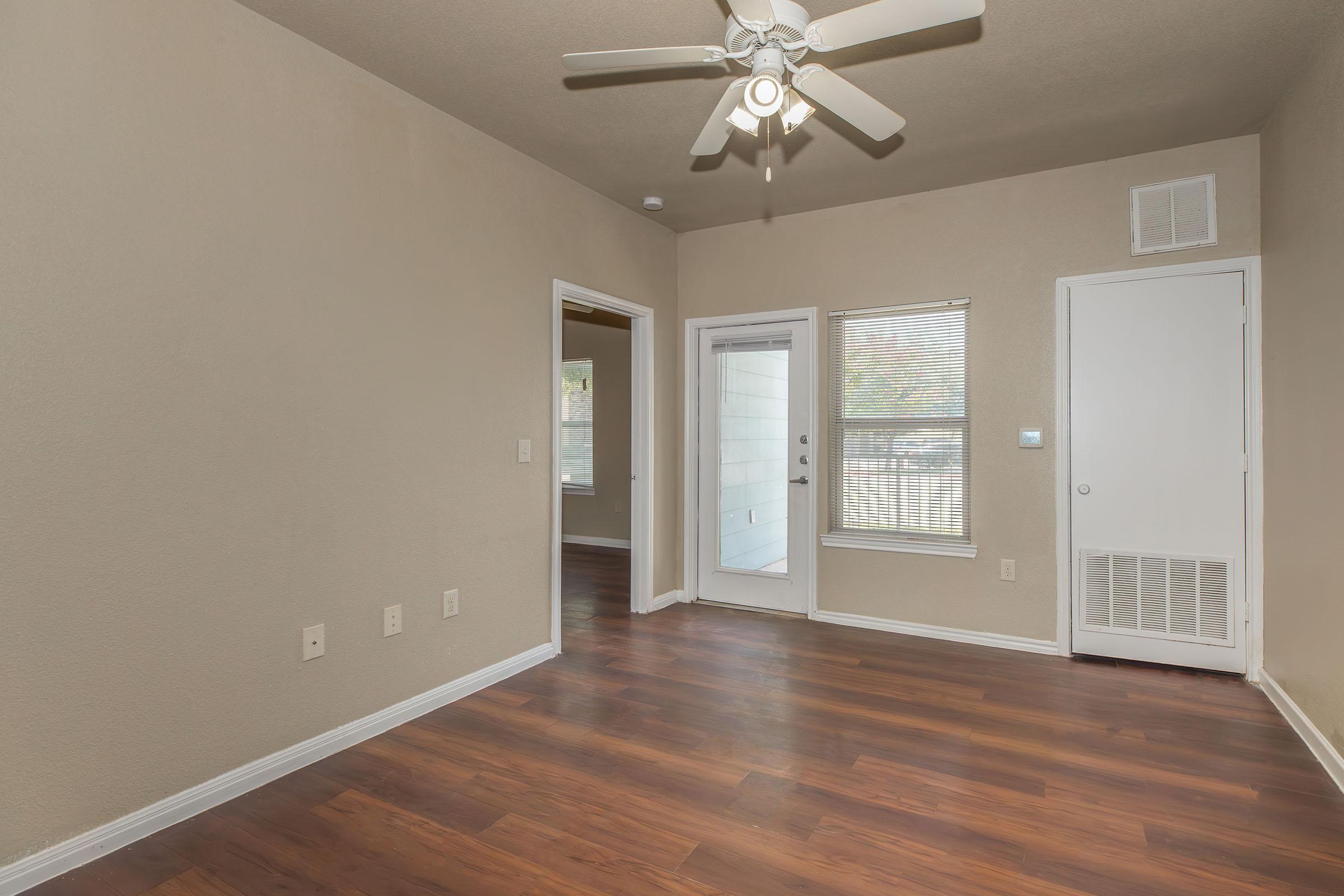 A bright, empty living room featuring light-colored walls and a ceiling fan. The floor is wooden and there are multiple entrances, including a door leading outside with a window beside it. A small door indicates a closet or storage area on the right.