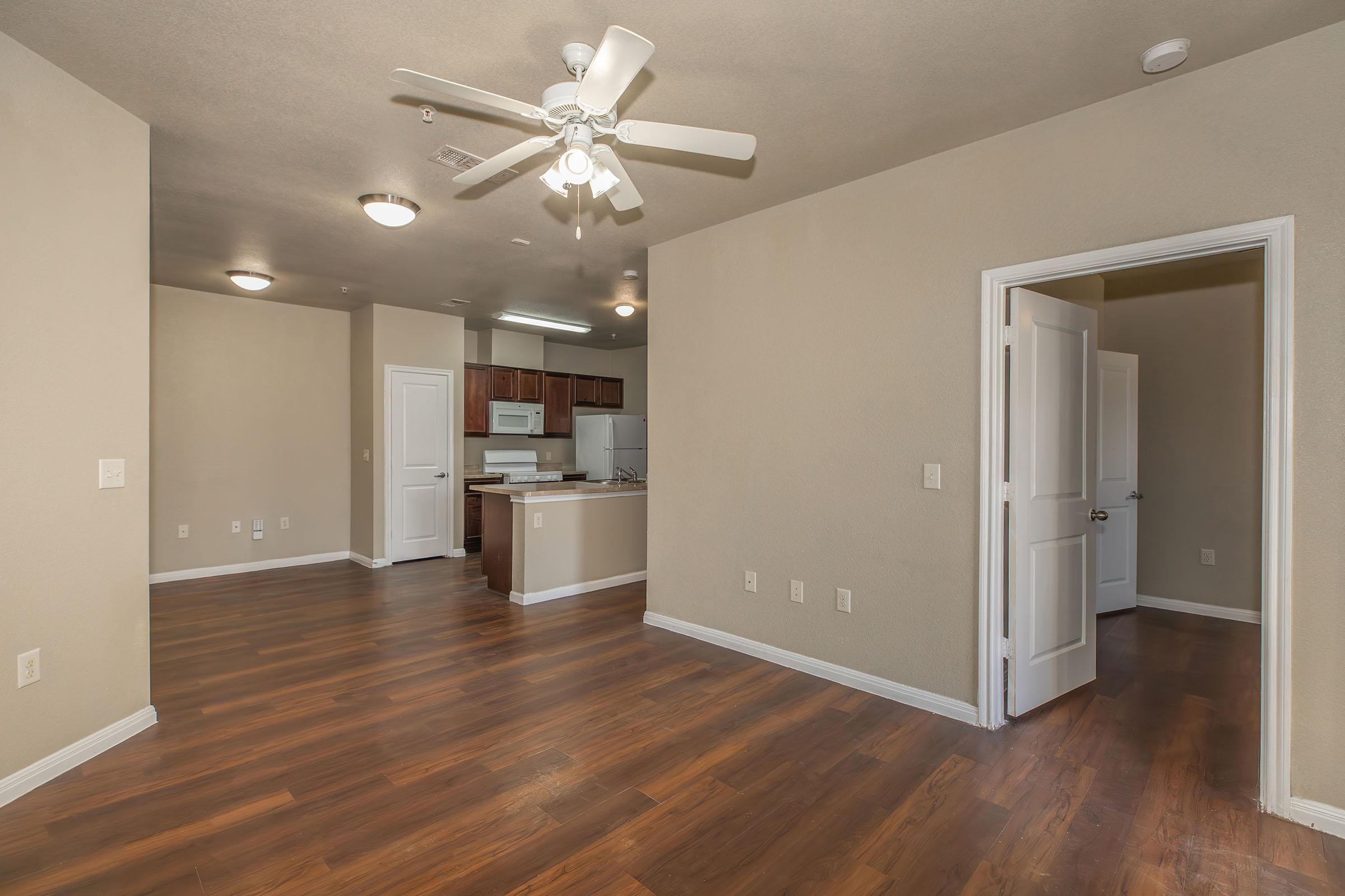 An interior view of a spacious, well-lit apartment featuring a living area with wood-style flooring, a ceiling fan, and light-colored walls. A doorway leads to a kitchen with modern appliances and cabinetry. The layout is open, giving a sense of flow between the living space and kitchen.
