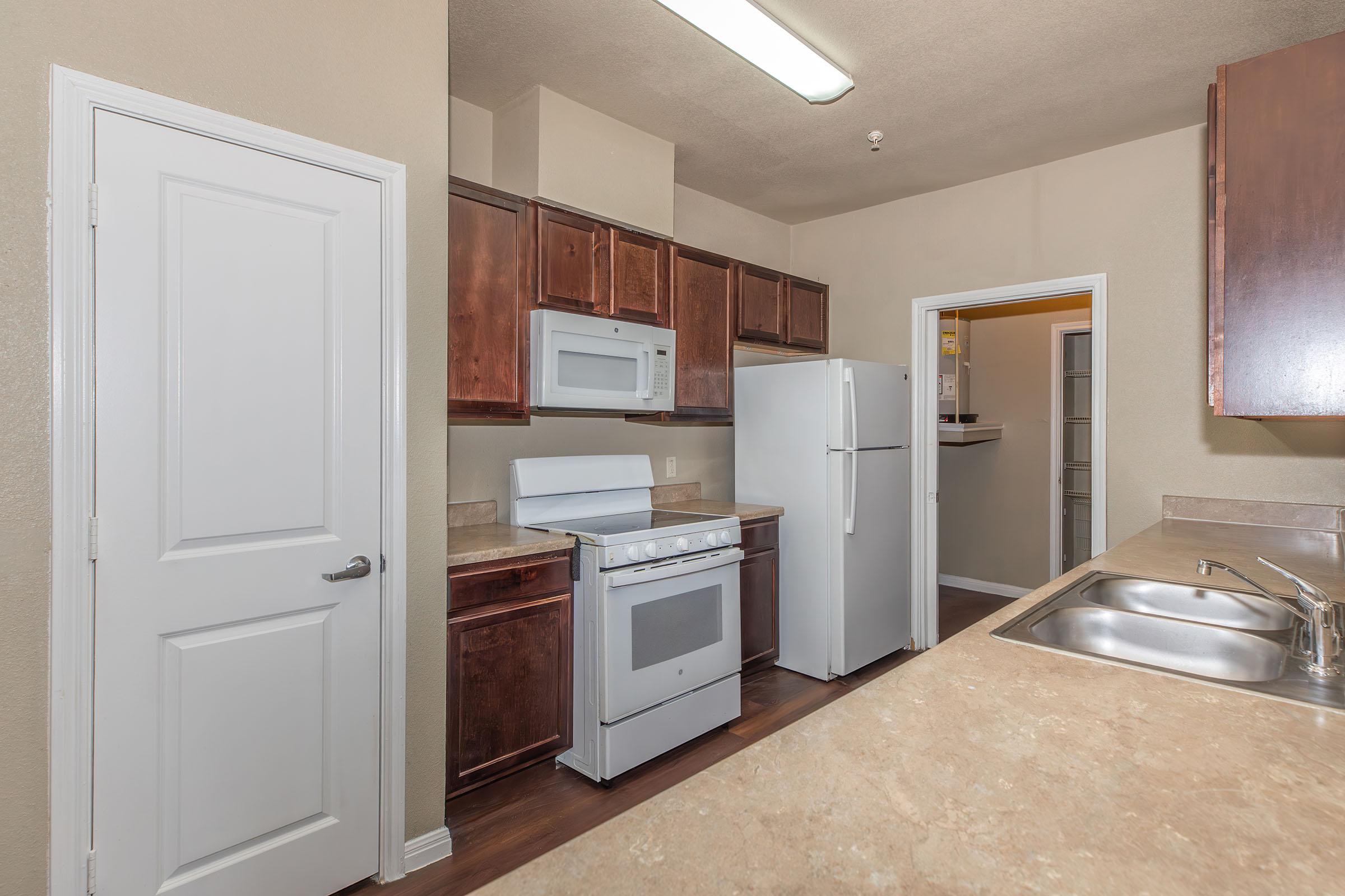 A modern kitchen featuring dark wood cabinets, a white stove, microwave, and refrigerator. The countertops are beige with a double sink visible. A doorway leads to another room, and the walls are painted in a light color. The kitchen is well-lit with overhead lighting.
