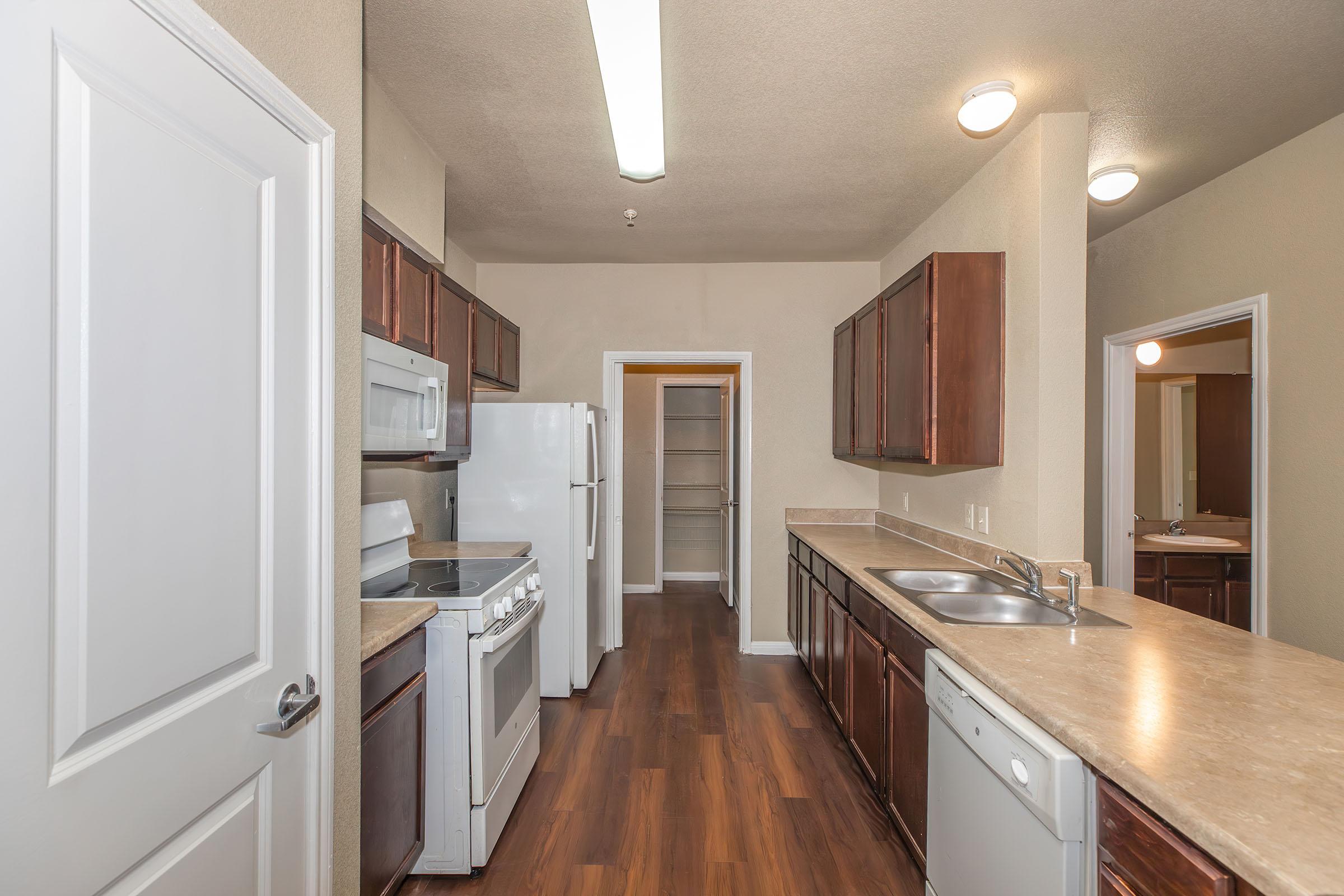 A spacious kitchen featuring wooden cabinetry, a white refrigerator, a gas stove, a microwave, and a dishwasher. The countertop is light-colored and the flooring is dark wood. There is a pantry visible through an open door at the end of the kitchen.