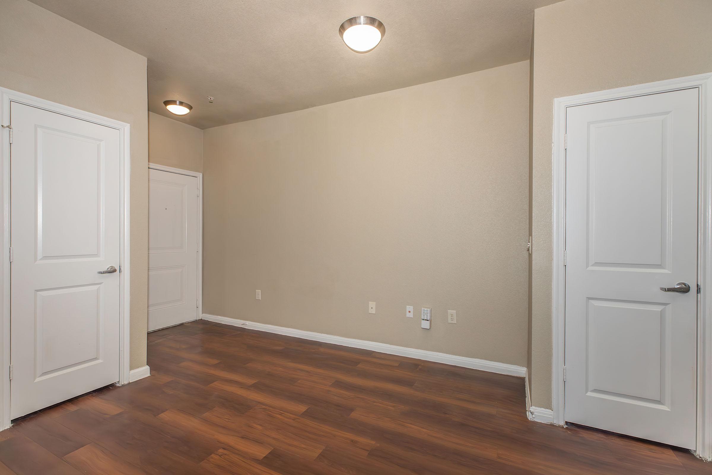 Empty room with light brown walls and a wooden floor. Two white doors are visible on the right side, and a door is located on the far left. The ceiling has two round light fixtures. The space appears well-lit and uncluttered, suitable for various uses.