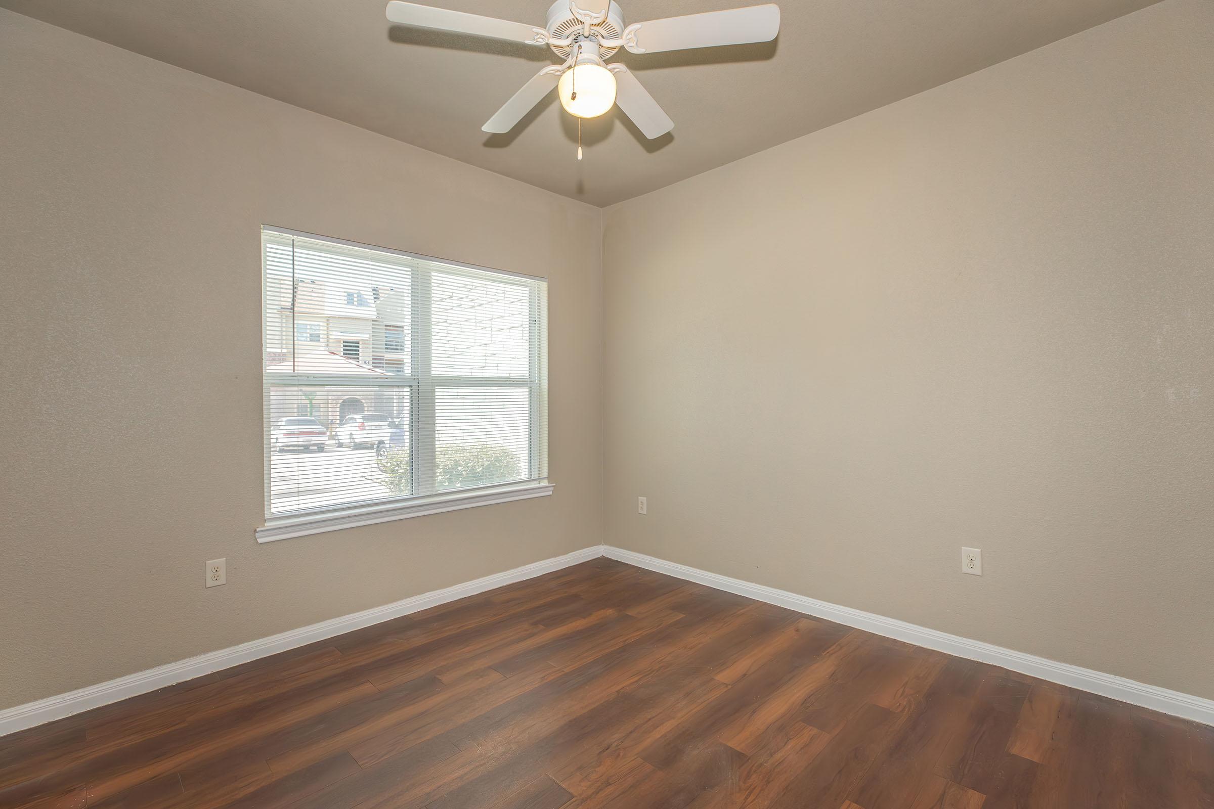 Empty room with light brown walls and a wooden floor. A ceiling fan is installed in the center, and natural light enters through a large window with white blinds. The view outside includes another building, but no furniture or decorations are present in the room.