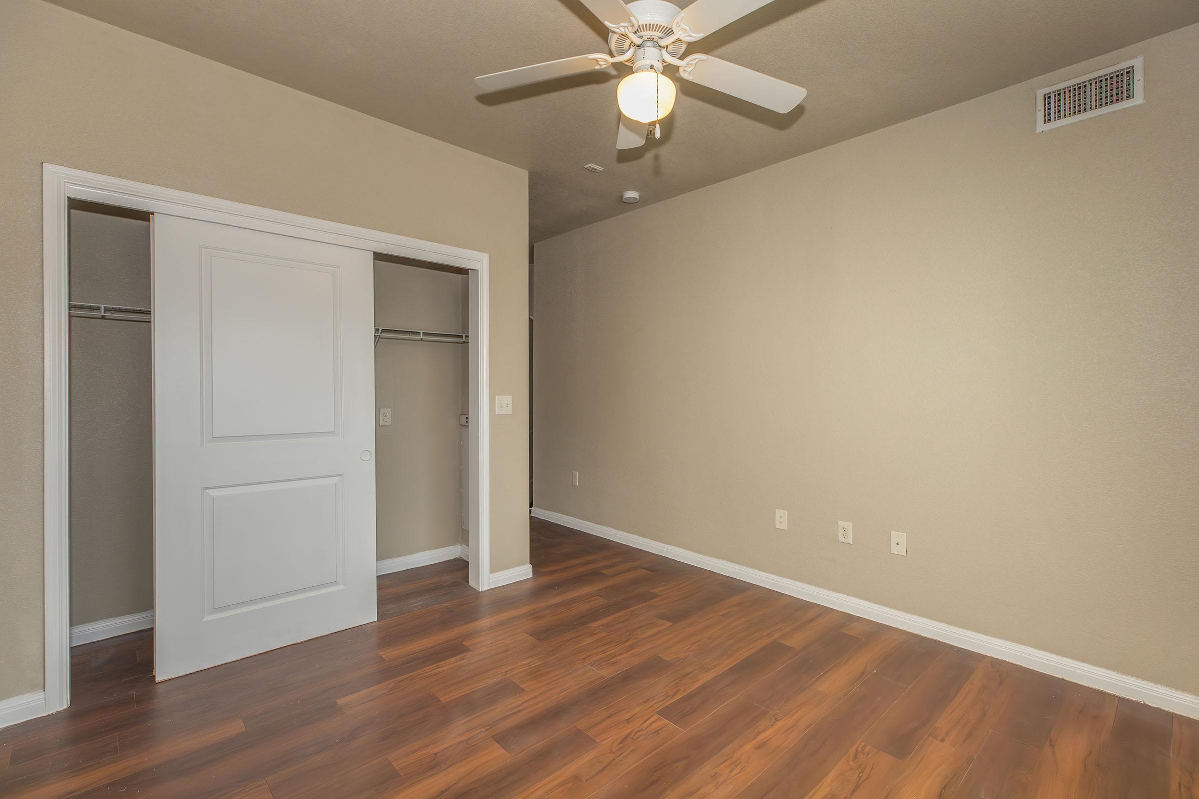 A spacious room with a light-colored wall and a wooden floor. It features a ceiling fan and a closed white door leading to a closet on the left. The room is empty, creating a clean, simple appearance, suitable for various uses. Natural light may come from the adjacent wall.