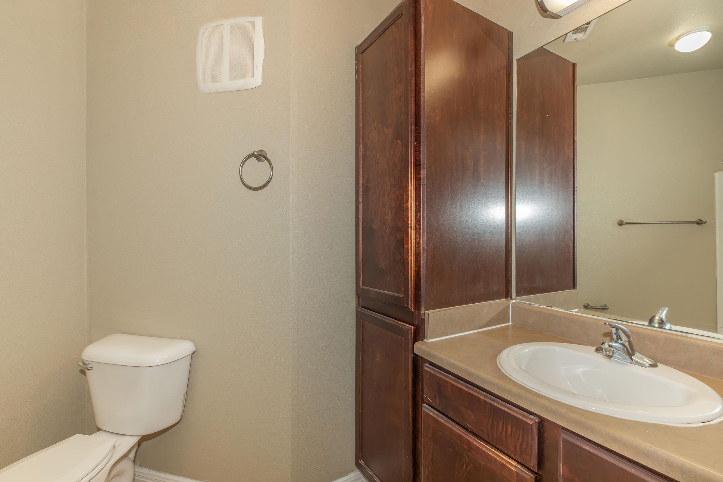 A clean bathroom featuring a toilet, a wooden vanity with a sink, and a mirror. The walls are painted a light color, and there is a towel rack visible. The overall design is modern and minimalist, with natural light coming from a ceiling fixture.