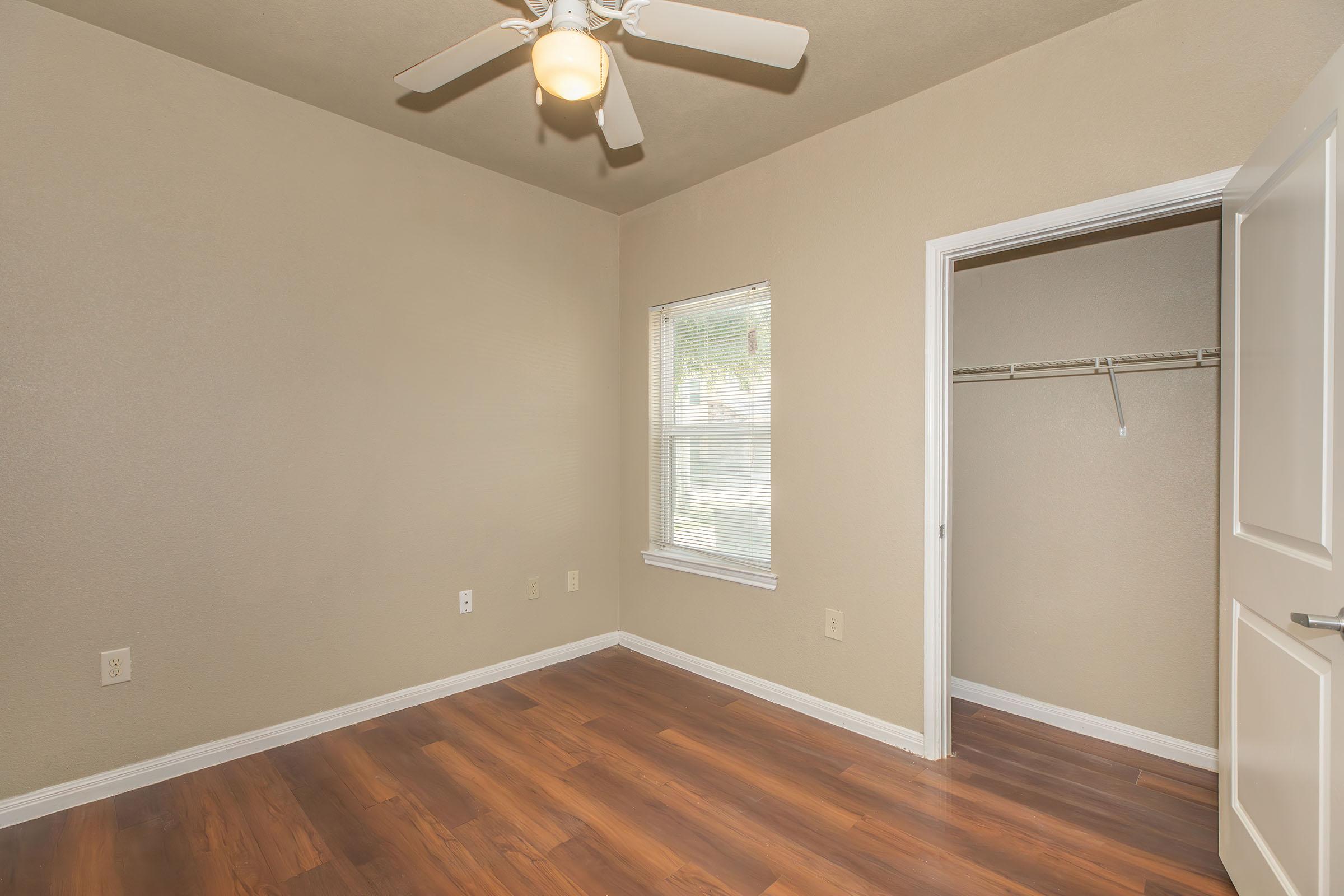 A small, empty room with beige walls and hardwood-style flooring. It features a ceiling fan, a window with natural light coming through, and an open closet with a hanging rod. The door to the closet is on the right side, while the window is positioned on the left wall.